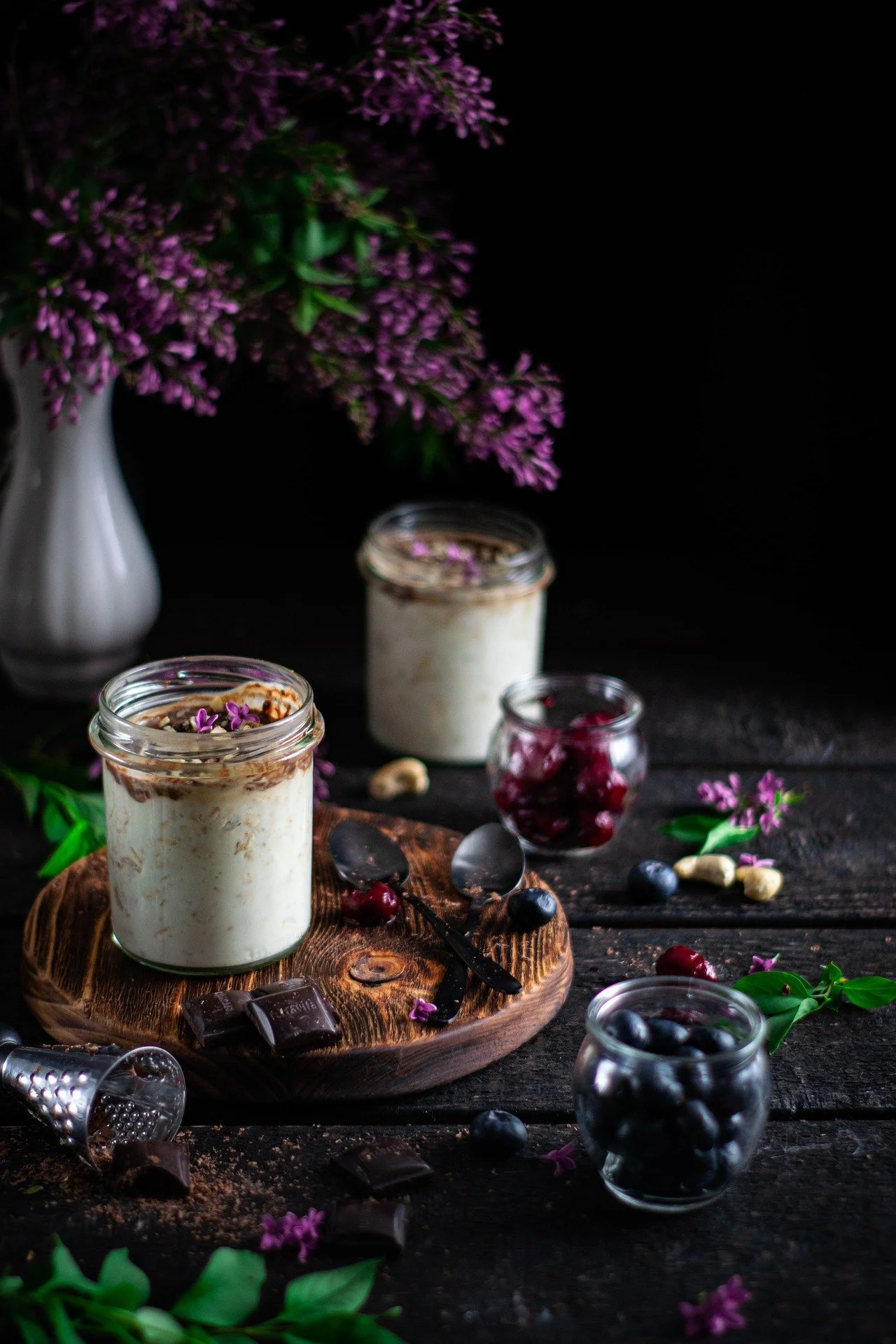 Two glasses of layered dessert with whipped cream, chocolate shavings, and purple flowers on a wooden tray, surrounded by blueberries, cherries, chocolate pieces, and small jars of berries on a rustic wooden table with purple flowers in a white vase 