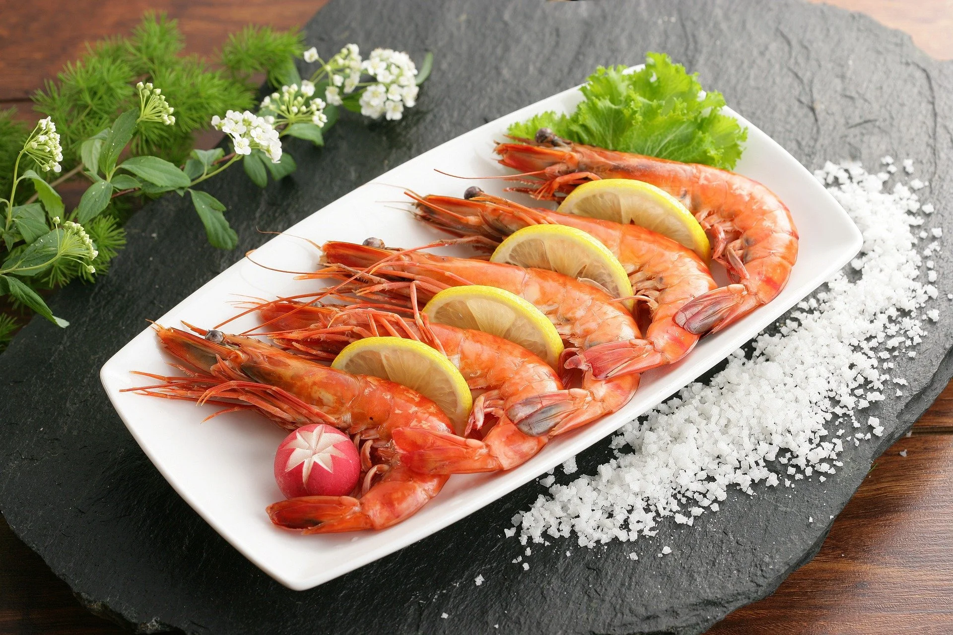 Fresh cooked shrimp garnished with lemon slices on a white platter, with lettuce, radish, and herbs as decoration, on a black slate serving board.