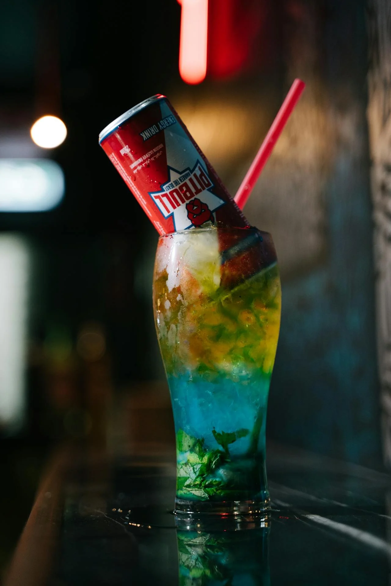 A colorful layered cocktail with mint leaves, ice, and a lime wedge in a tall glass. A can of Red Bull energy drink is inserted upside down into the drink, and a pink straw protrudes from the glass.