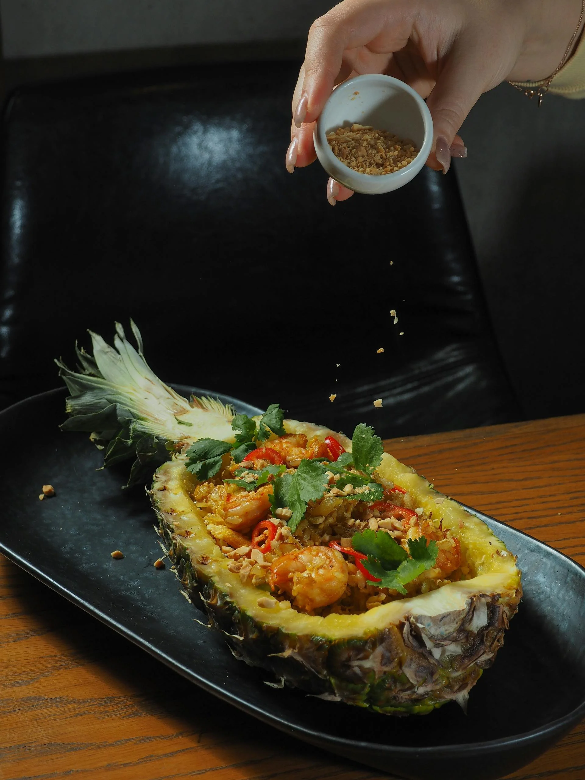 Pineapple boat filled with shrimp, rice, red chili slices, cilantro, and crushed peanuts on a black plate, with a hand sprinkling crushed peanuts from a small white bowl.