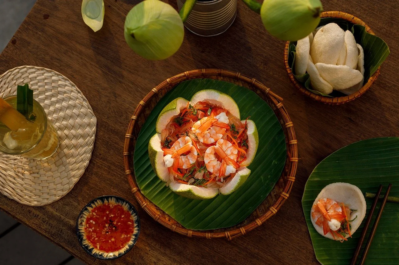 A seafood dish with shrimp, herbs, and vegetables served on a banana leaf on a wicker tray, accompanied by lime slices, rice crackers, a dipping sauce, and a drink with a straw, all on a wooden table.