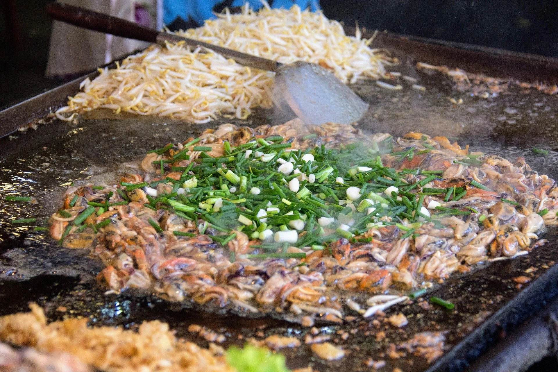 Cooking food on a large flat griddle, including chopped green onions and shellfish, with noodles in the background.