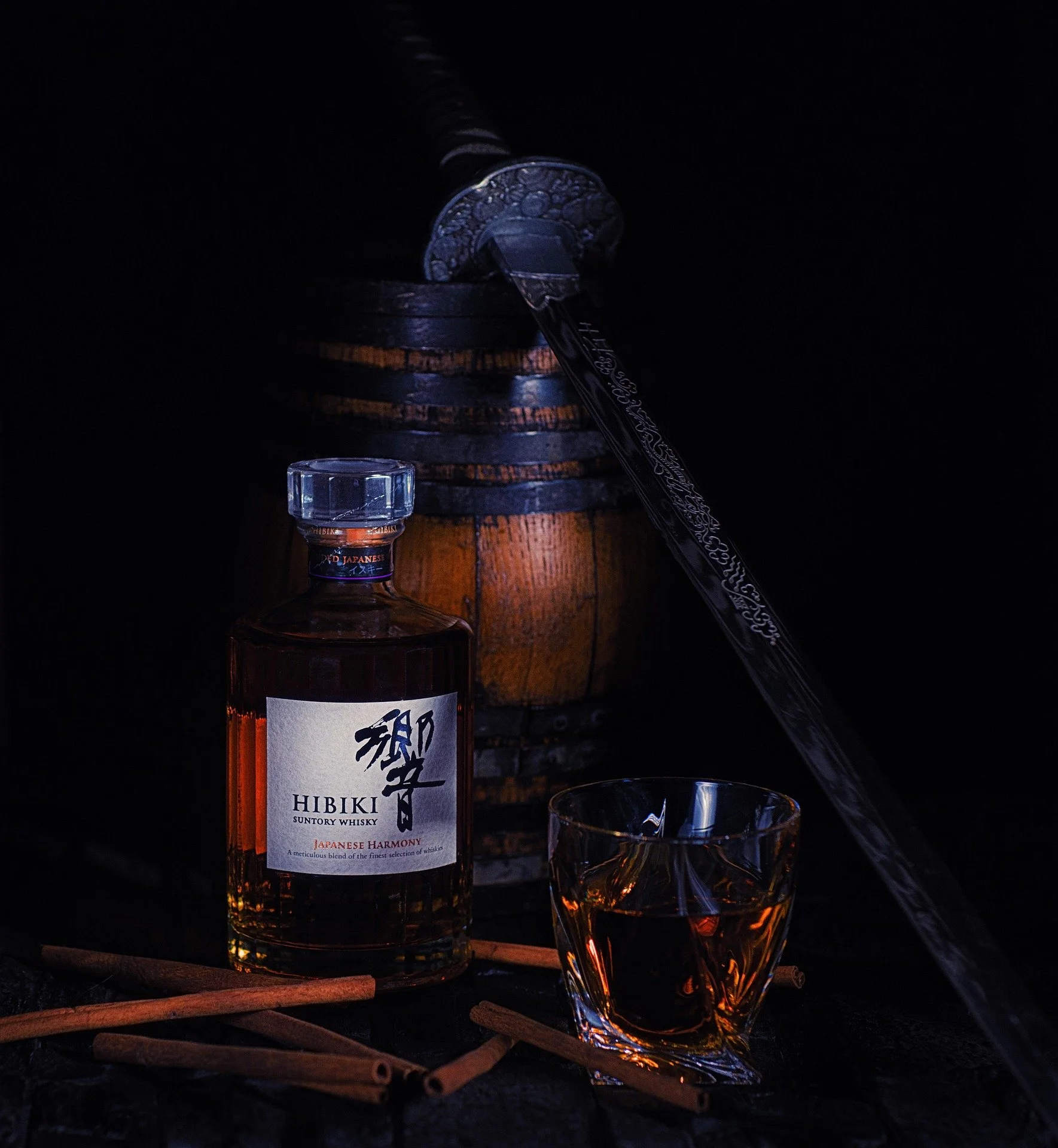 A bottle of Hibiki Japanese Harmony whisky, a glass of whisky, cinnamon sticks, a Japanese sword, and a wooden barrel against a dark background.
