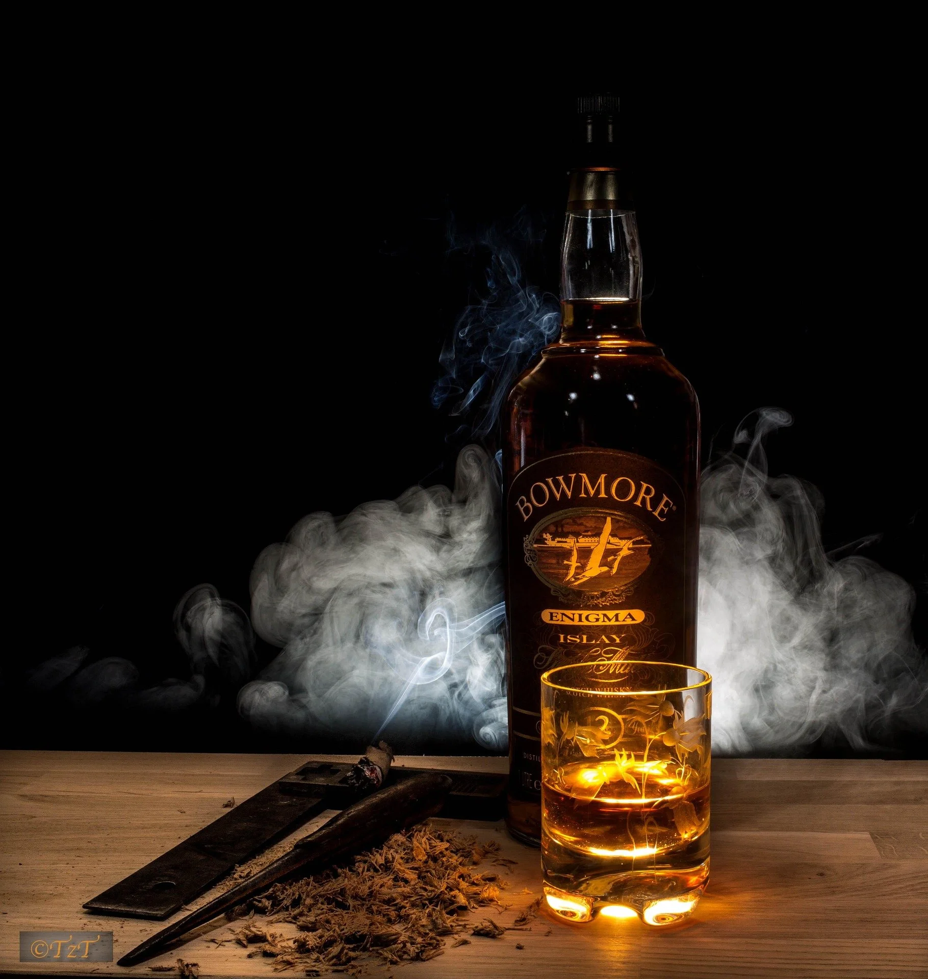 A bottle of Bowmore Enigma Islay Scotch whisky, a glass filled with whisky, a lit cigar resting on a wooden surface with ash and a cigar cutter, and smoke swirling around on a dark background.