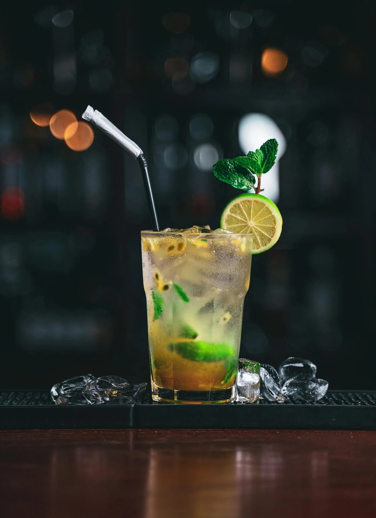 A glass of cocktail with lime, mint leaves, ice, and a straw on a bar counter with ice cubes around it.