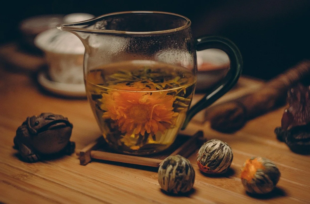 Glass teapot with a yellow flower inside, placed on a wooden surface with tea leaves, nuts, a wooden spoon, and a metal tray in the background.