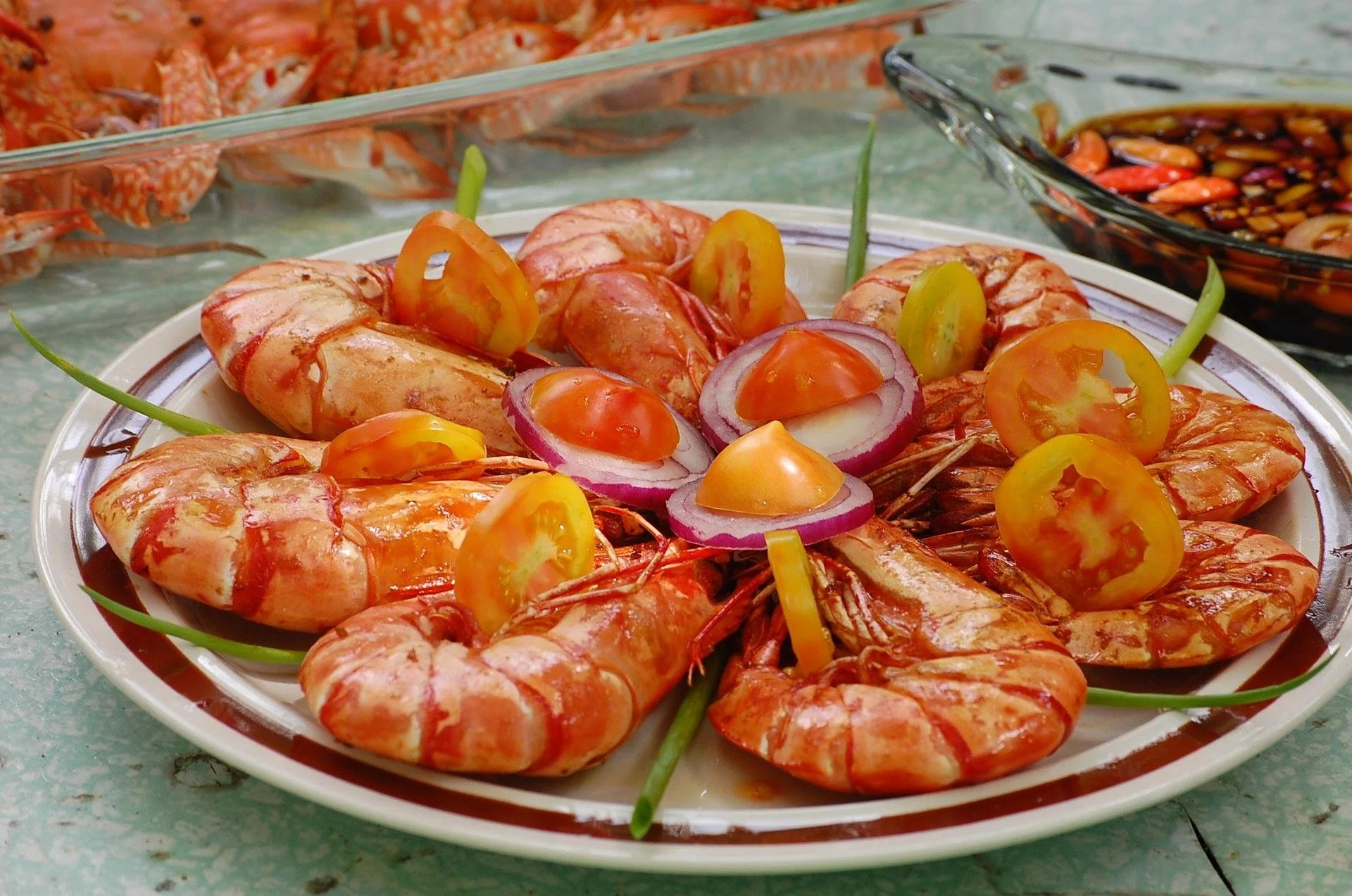 Plate of cooked shrimp garnished with sliced yellow and red tomatoes, red onion slices, and green onions, with a bowl of mixed seafood in the background.