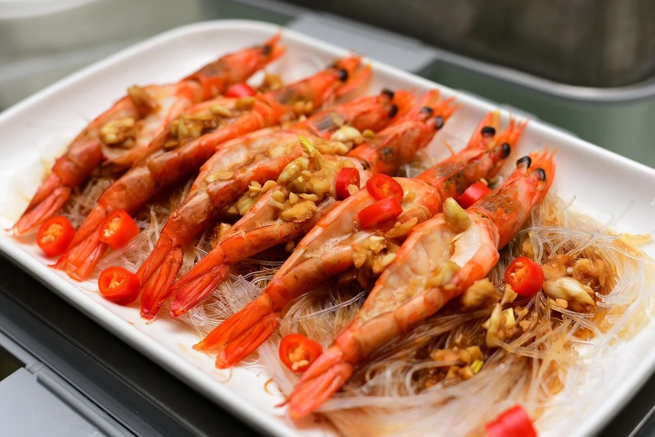 A platter of cooked shrimp topped with chopped garlic and red chili slices, served on a bed of vermicelli noodles.