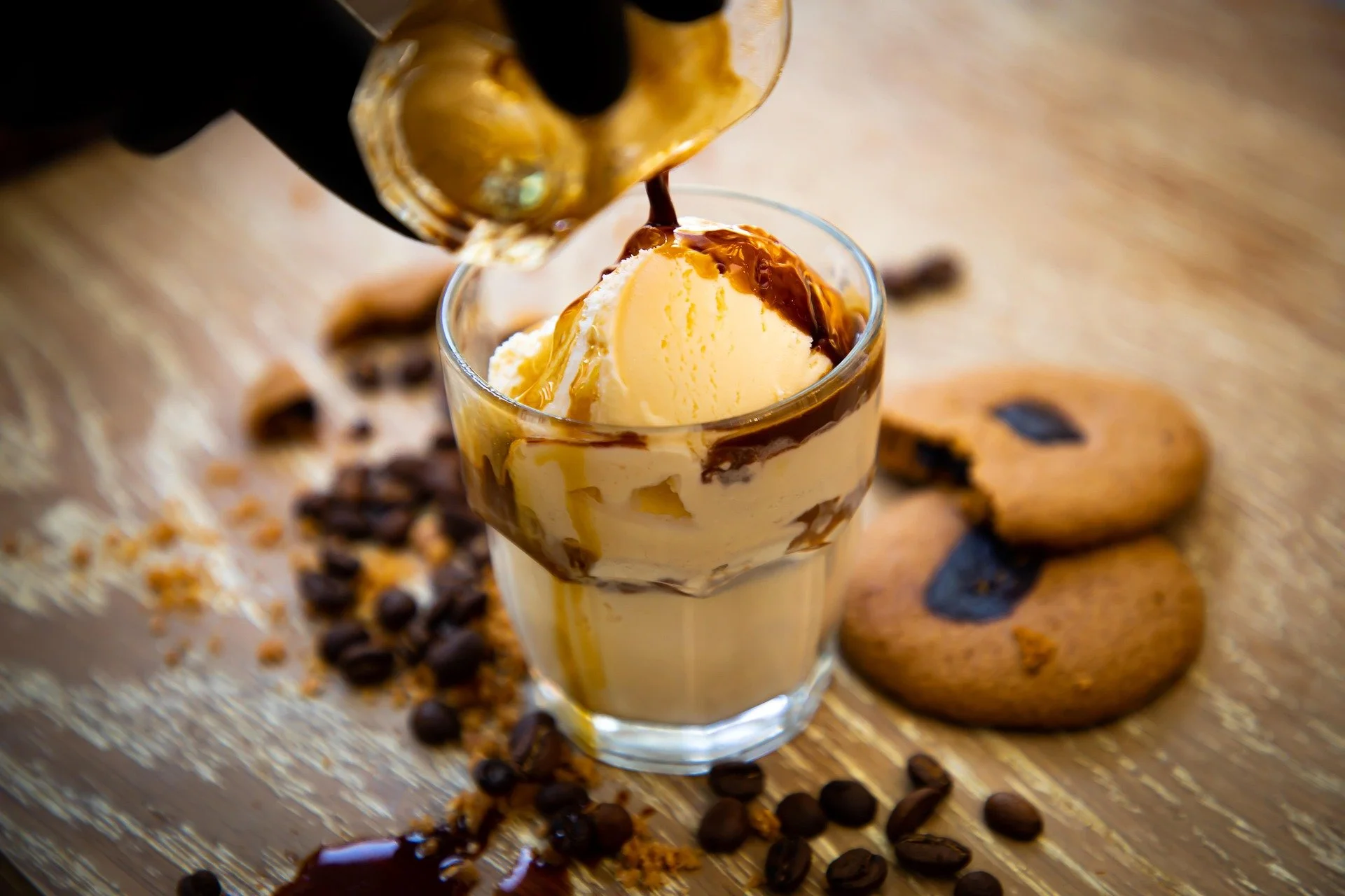 A glass of vanilla ice cream topped with caramel sauce, with chocolate cookies and coffee beans scattered on a wooden surface in the background.