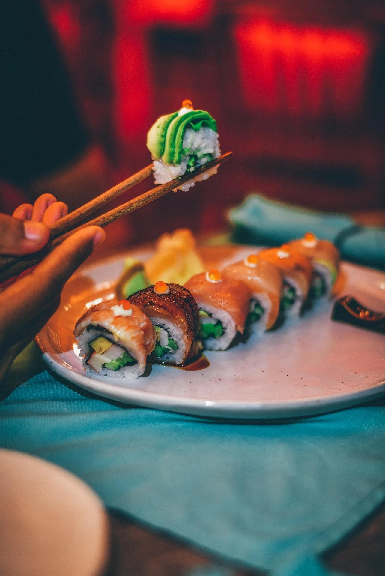 Close-up of a plate of assorted sushi rolls on a table, with a person using chopsticks to pick up a sushi piece topped with green and white icing or decoration. The background has warm red lighting.