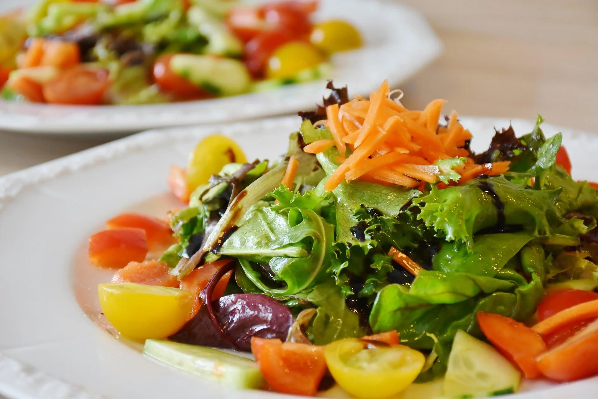 Fresh mixed green salad with shredded carrots, cherry tomatoes, cucumber slices, and dark balsamic dressing on a white plate.