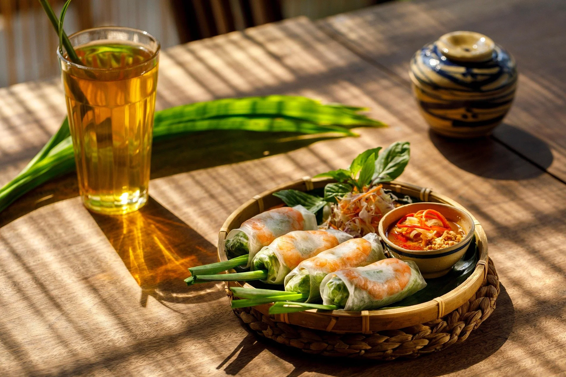 Spring rolls served with fresh herbs and dipping sauce on a woven tray, alongside a glass of iced tea with a straw, on a wooden table with sunlight and shadows.