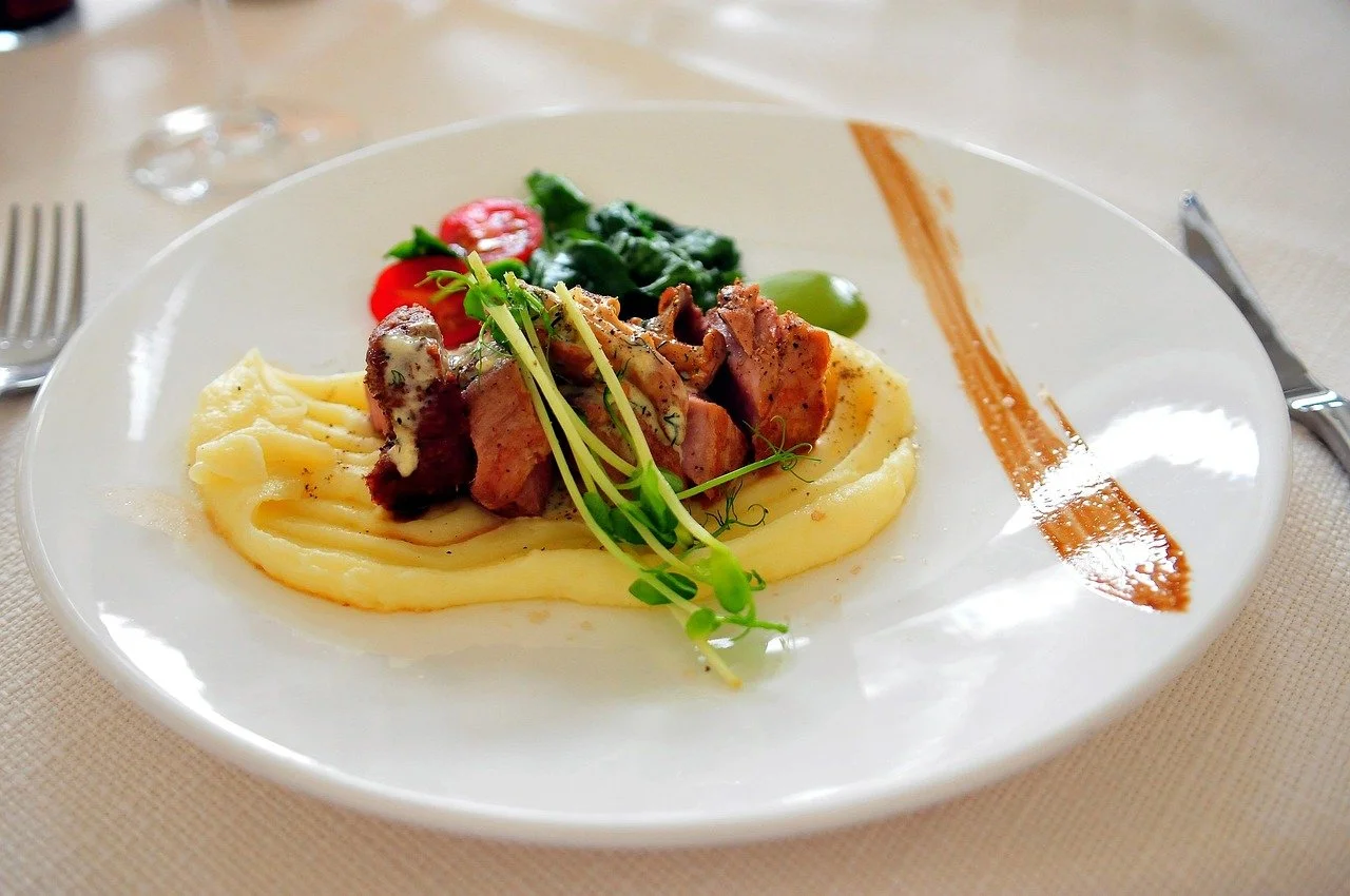 A plated gourmet dish featuring mashed potatoes topped with slices of cooked meat, garnished with microgreens, and set on a white plate with a decorative sauce stripe. There are also cherry tomato, spinach, and green olive in the background.