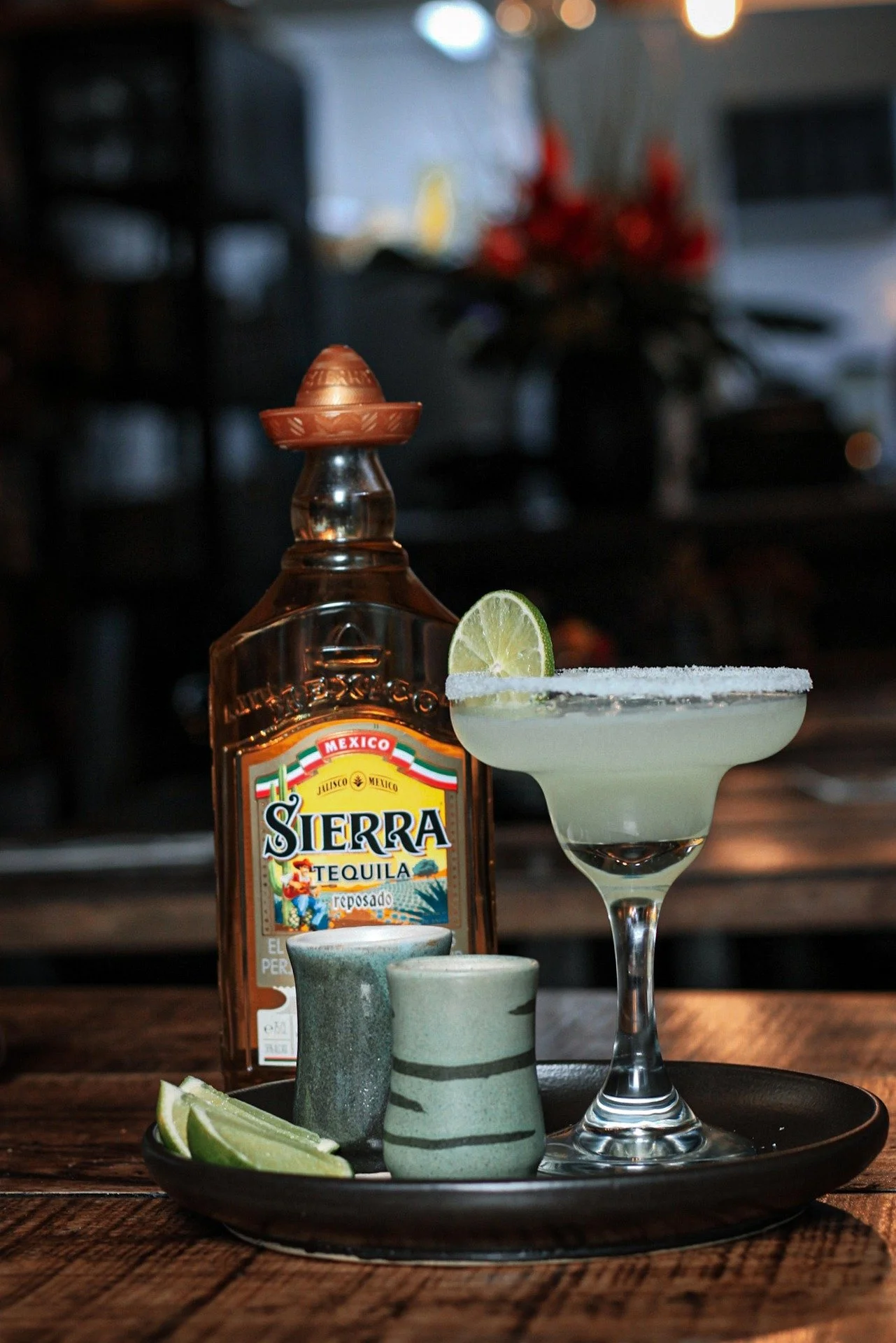 A margarita cocktail with lime wedge garnishing, Sierra tequila bottle, two small cups, and lime slices on a black plate on a wooden table.