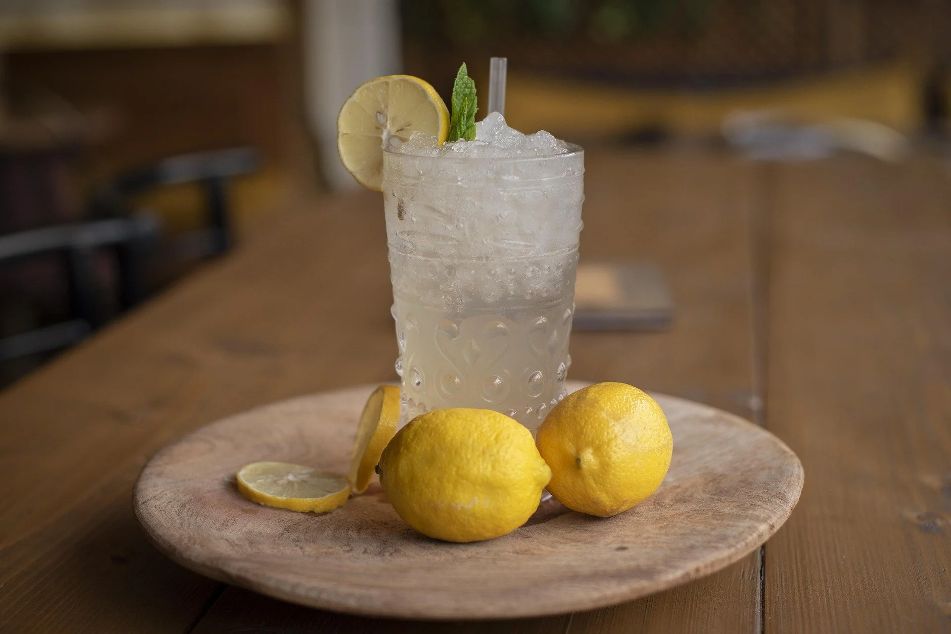 A tall glass of lemonade with ice, garnished with a lemon slice, mint, and a straw, placed on a round wooden plate with whole and sliced lemons around it, on a wooden table.