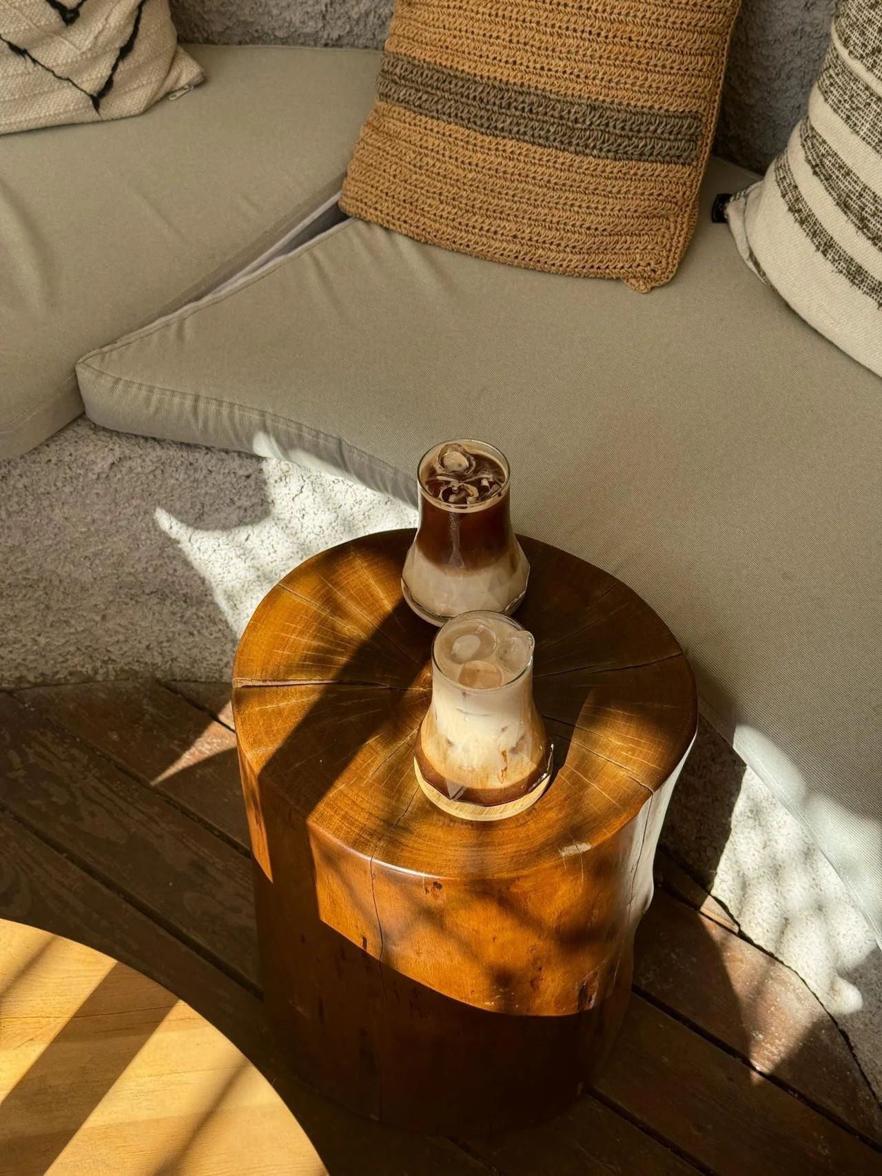 Two iced coffee drinks with cream and ice on a round wooden side table next to a beige sofa with patterned pillows.