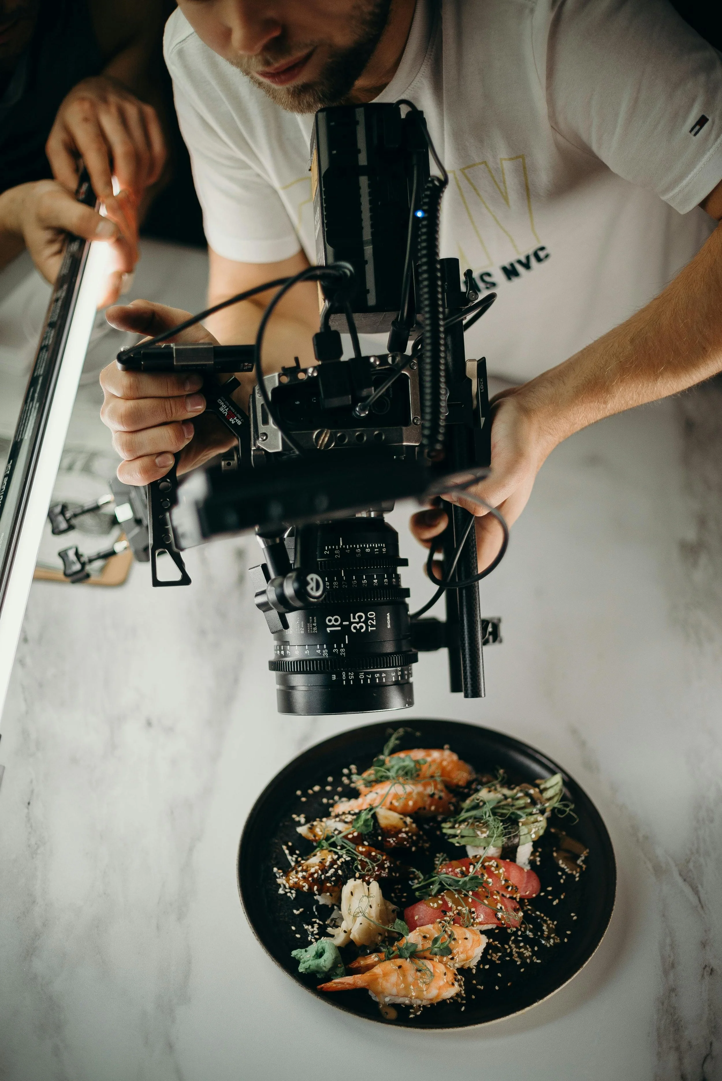 A photographer takes close-up shots of a plated dish with shrimp, vegetables, and garnishes using a professional camera mounted on a stabilizer.