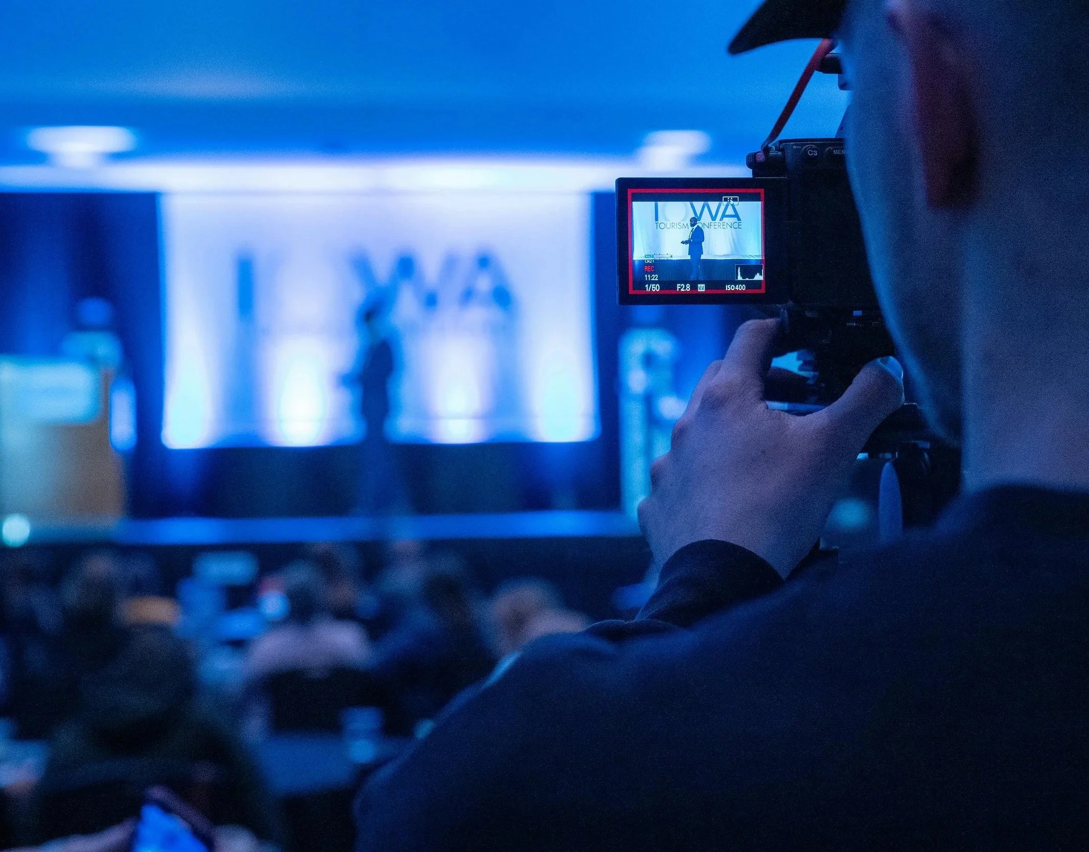 A person is filming a speaker on stage at a conference. The stage has a large backdrop that reads 'WWA.' The scene is illuminated with blue lighting, and the photo is taken through a camera viewfinder.