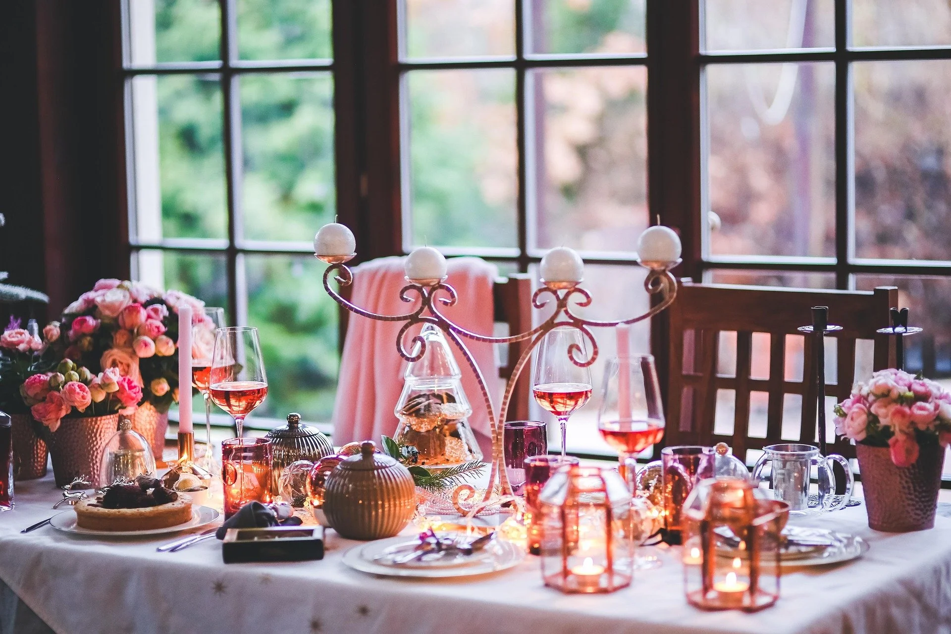A dining table decorated with pink flowers, candles, wine glasses with rosé wine, and a candelabra, set near large windows with a view of greenery outside.