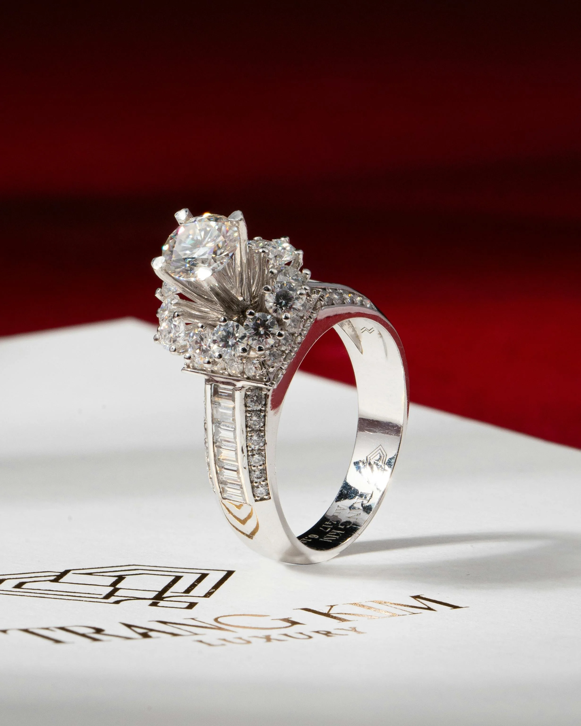A silver engagement ring with multiple diamonds, featuring a large central diamond surrounded by smaller diamonds, placed on a white cloth with a red background.