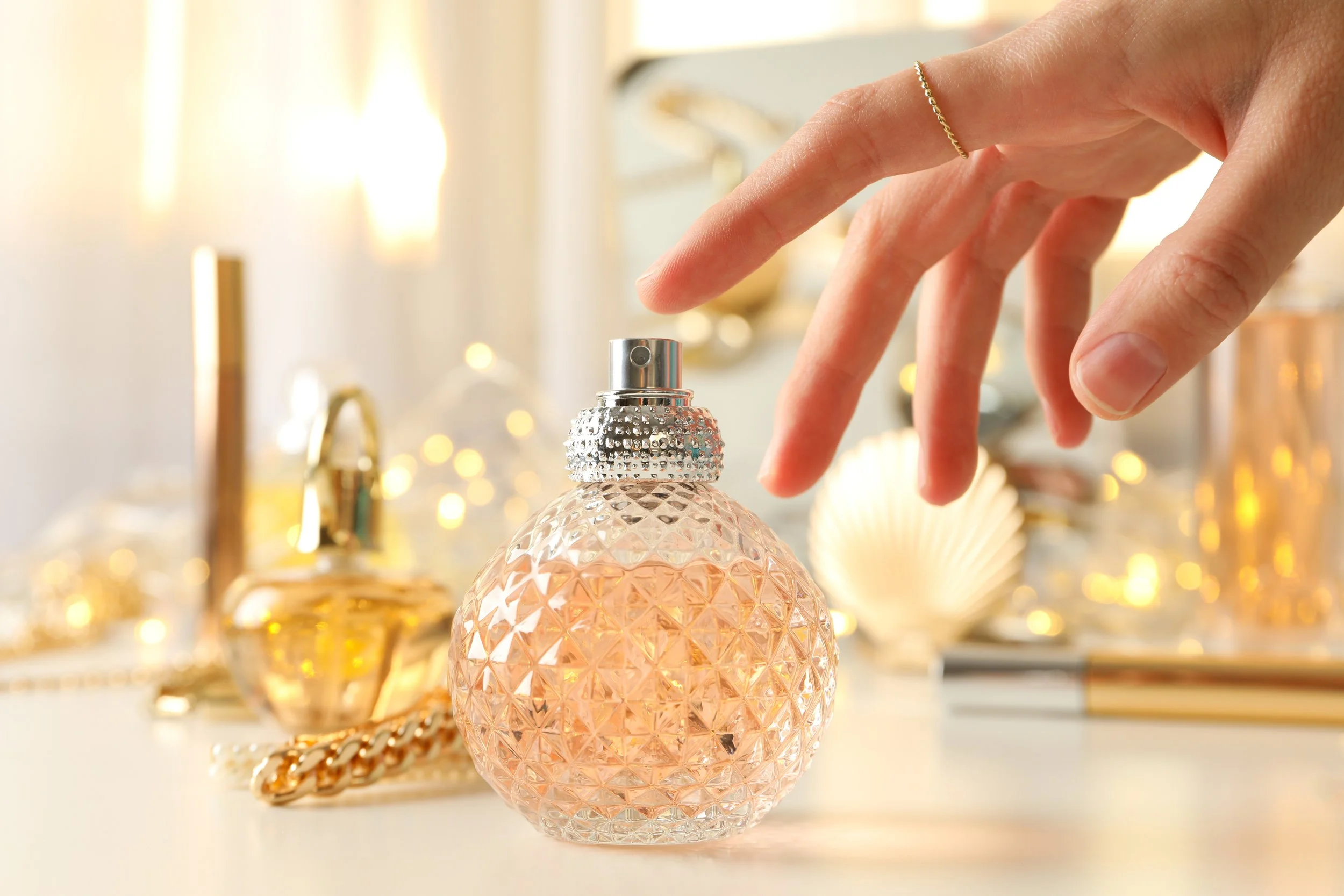 Hand pressing perfume atomizer on a glass perfume bottle with a decorative background of gold items and lights