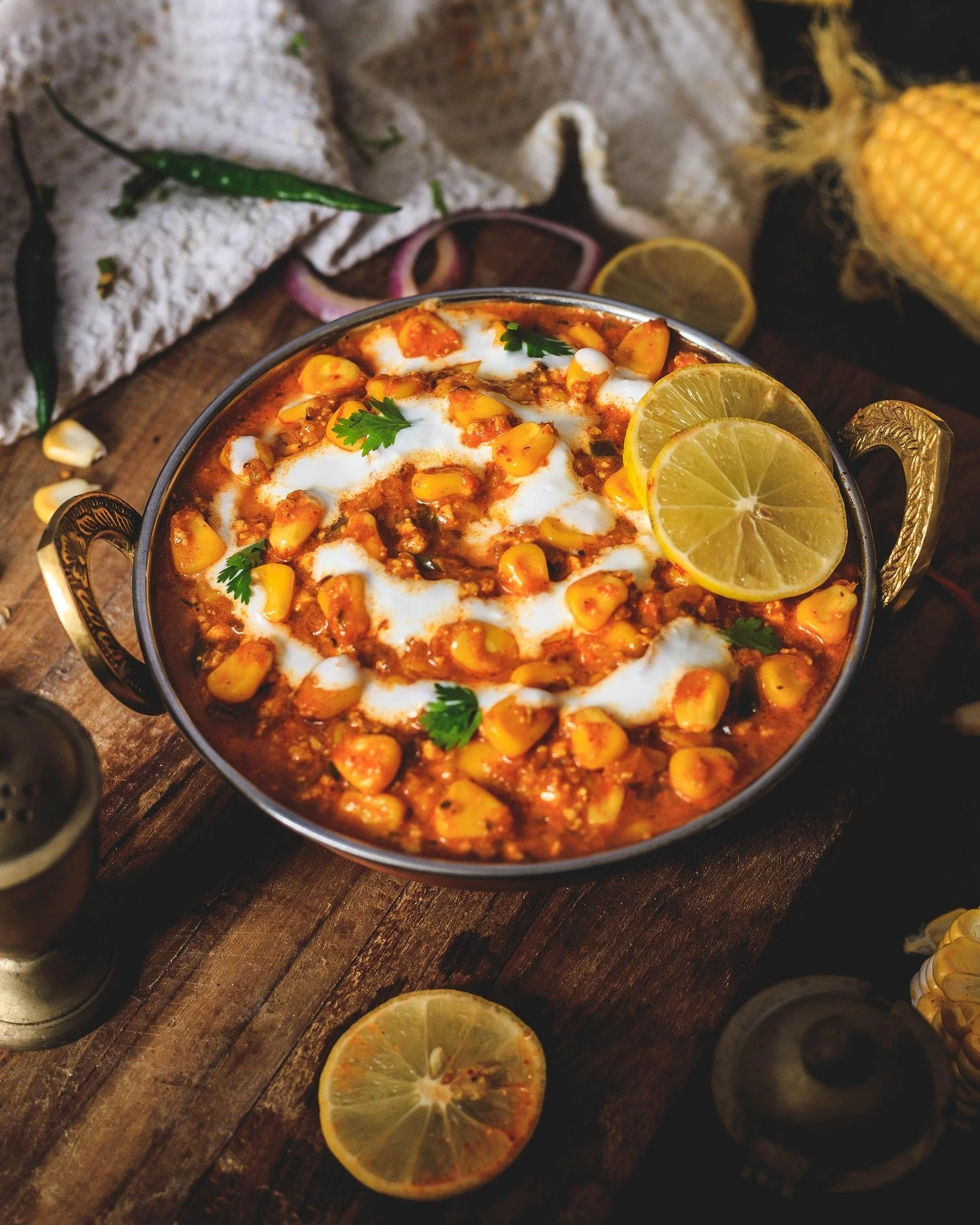 A bowl of Indian chickpea curry garnished with lemon slices, cilantro, and yogurt, placed on a rustic wooden table surrounded by lemon slices, corn, and spices.