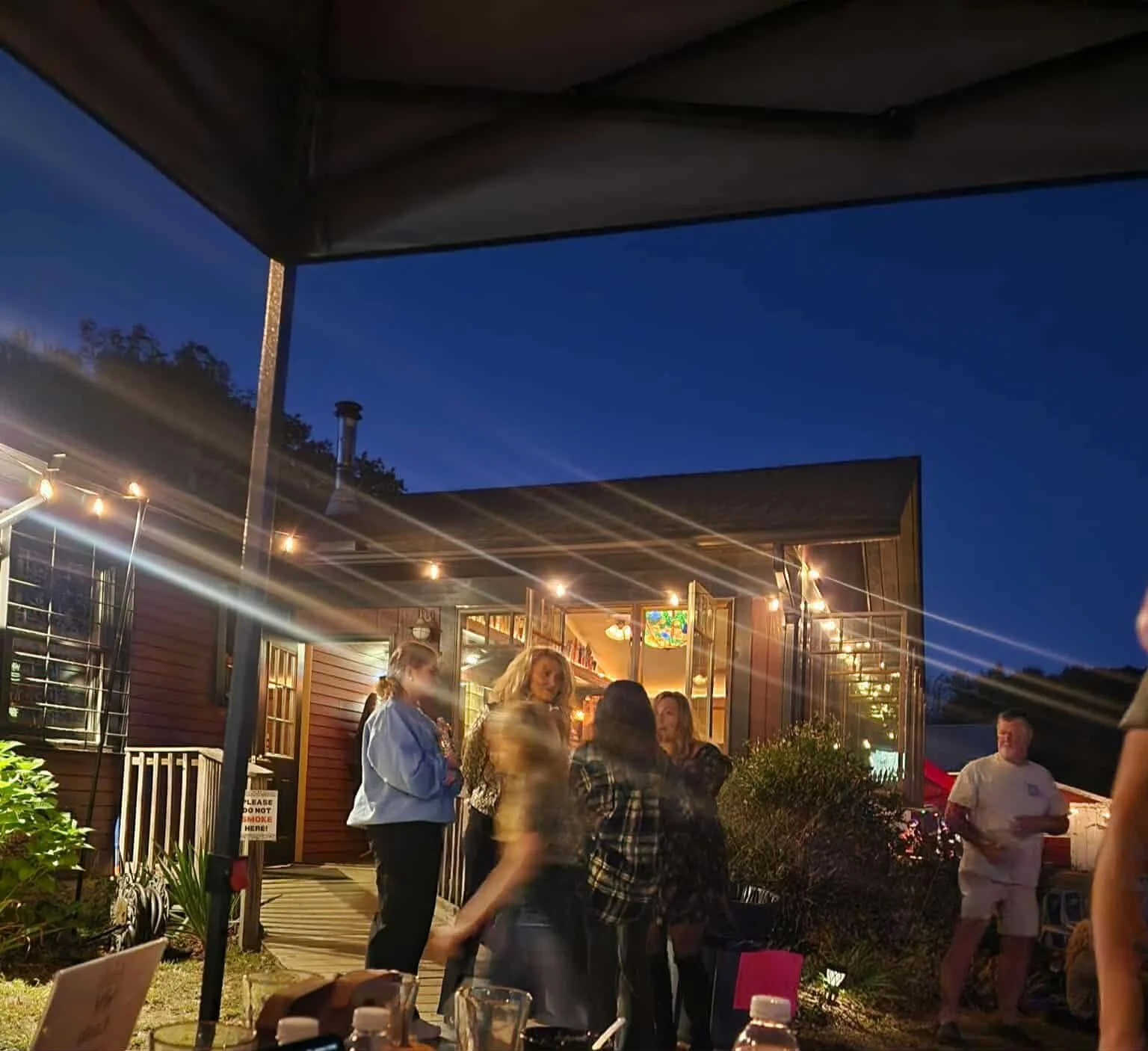 Group of people gathered outside a restaurant at night, illuminated by string lights, with a blue sky in the background.