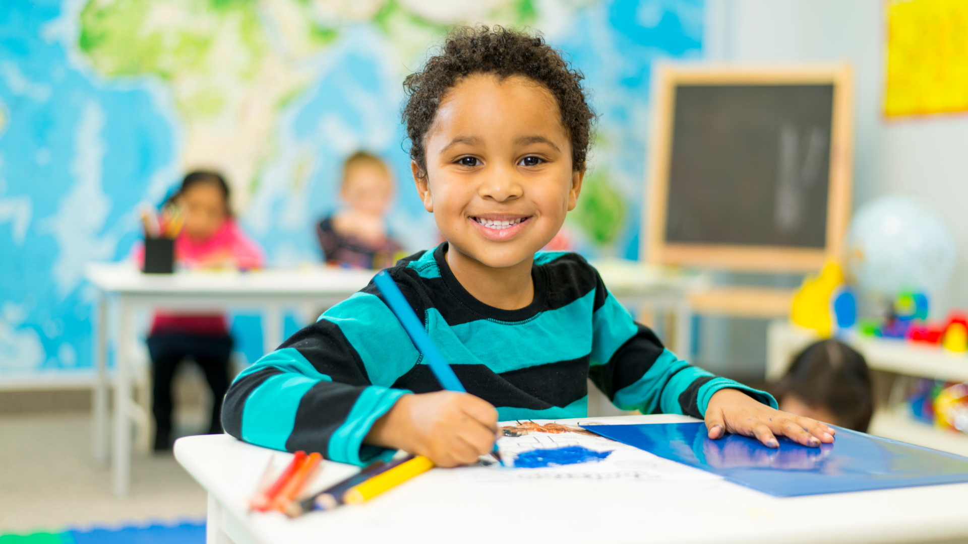 Smiling young girl drawing with colored markers at a table in a colorful classroom with a world map background.