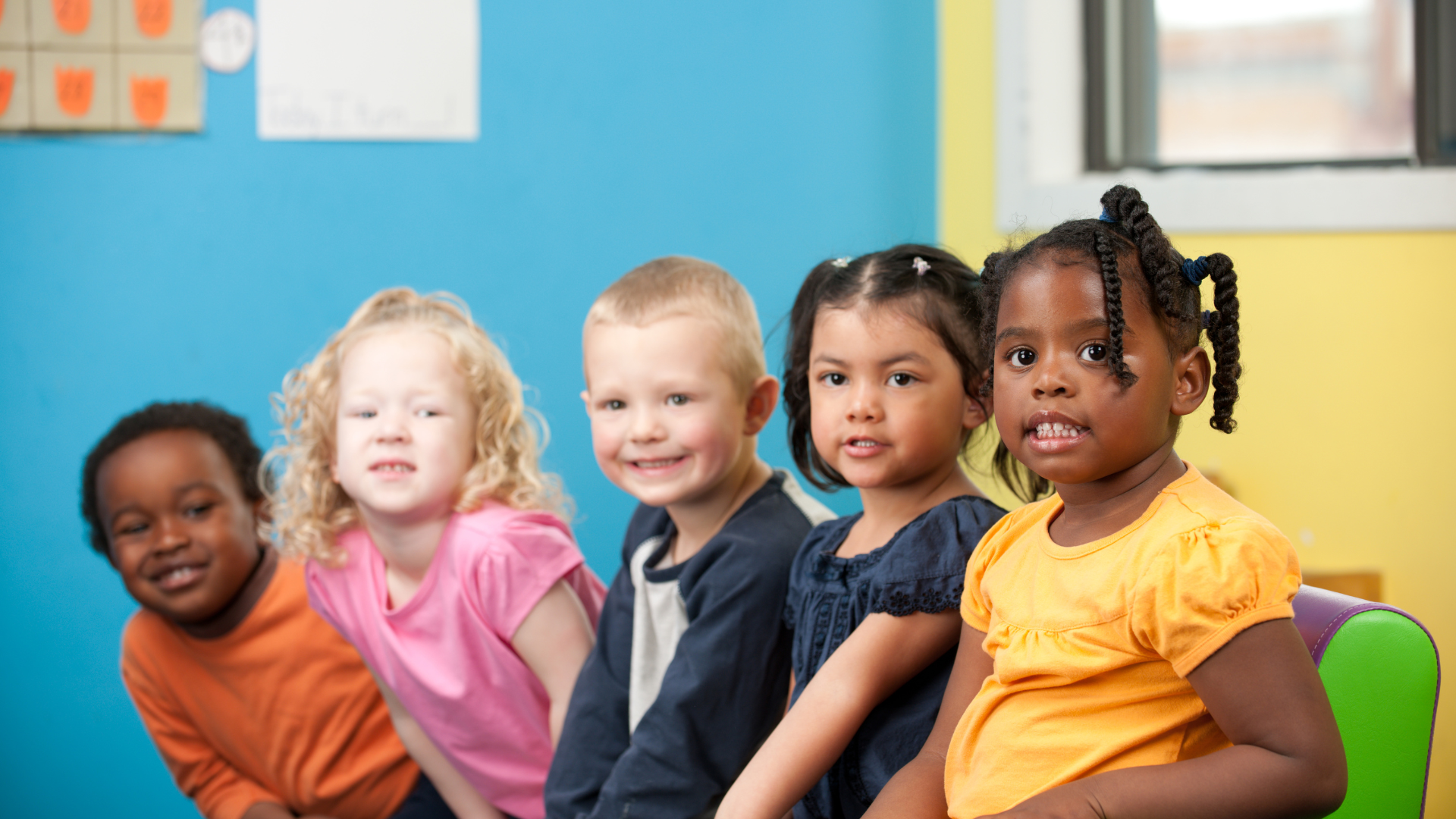 Five children sitting in a row in a classroom, smiling and looking at the camera.