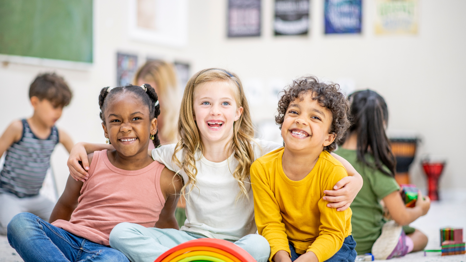 Three smiling children sitting together in a classroom, with other children in the background, and toys and a rainbow in the foreground.