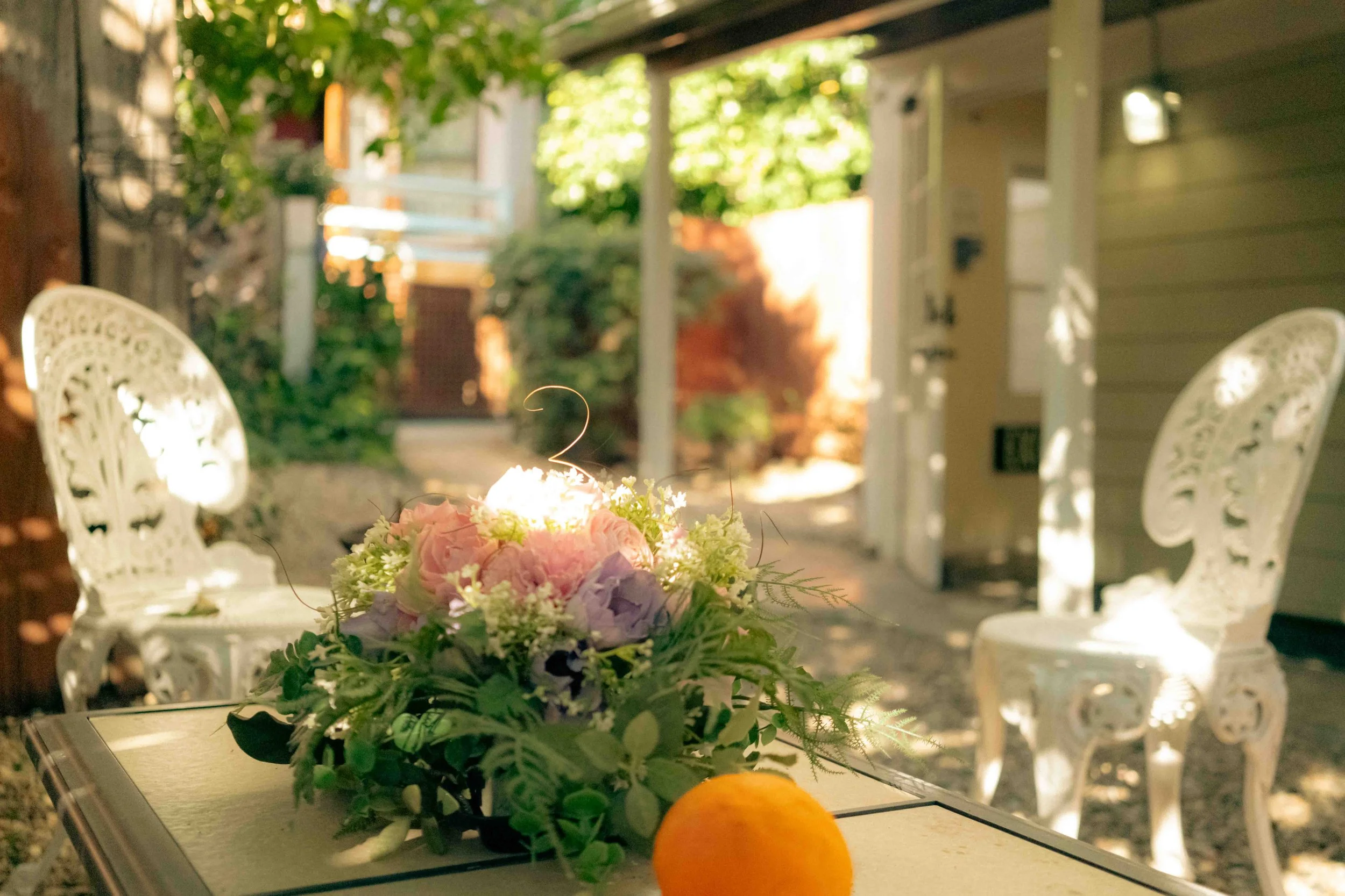 Outdoor patio with two ornate white chairs, a small table with a floral centerpiece and an orange, surrounded by greenery and dappled sunlight.