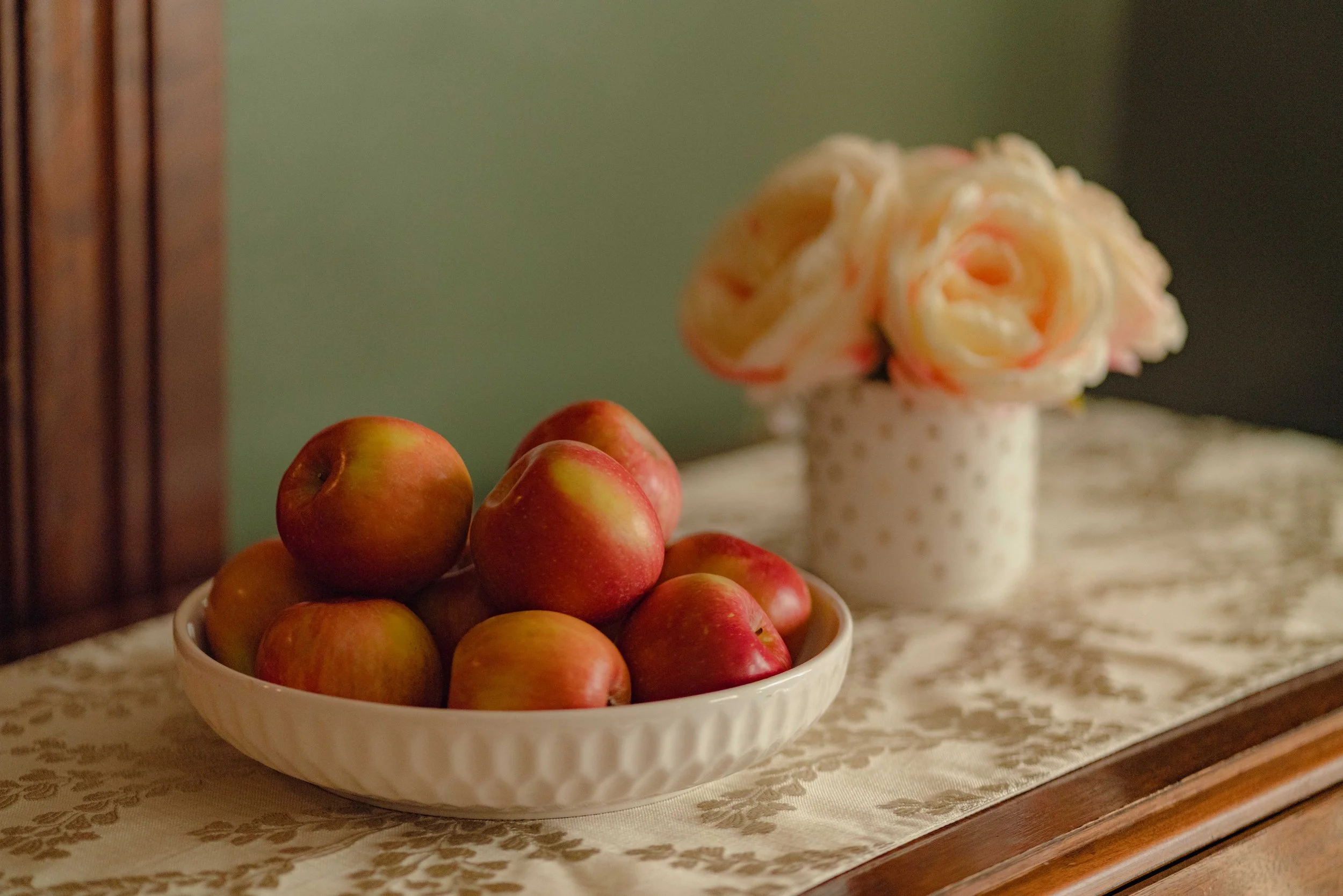 Bowl of red apples on a table with a vase of pink roses