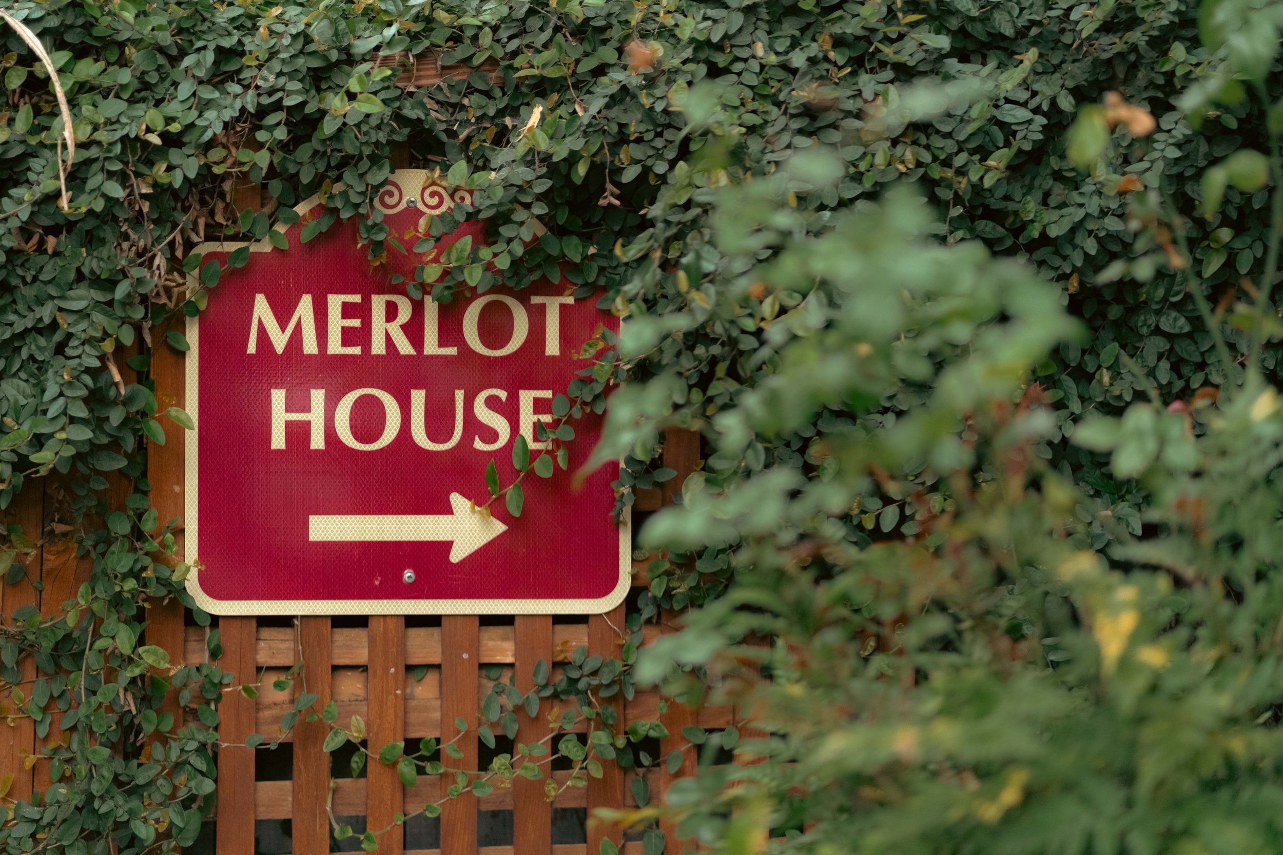 A wooden sign reading 'Merlot House' with an arrow pointing right, surrounded by lush green foliage.