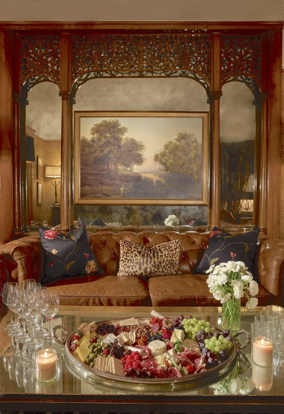 A cozy living room with a leather tufted sofa decorated with floral and leopard print pillows. A glass coffee table with a tray of assorted cheeses, fruits, and desserts. A flower arrangement in a glass vase and four candles on the table. A large landscape painting hangs on a wall with decorative woodwork above the mirror, reflecting the warm lighting and part of the room.
