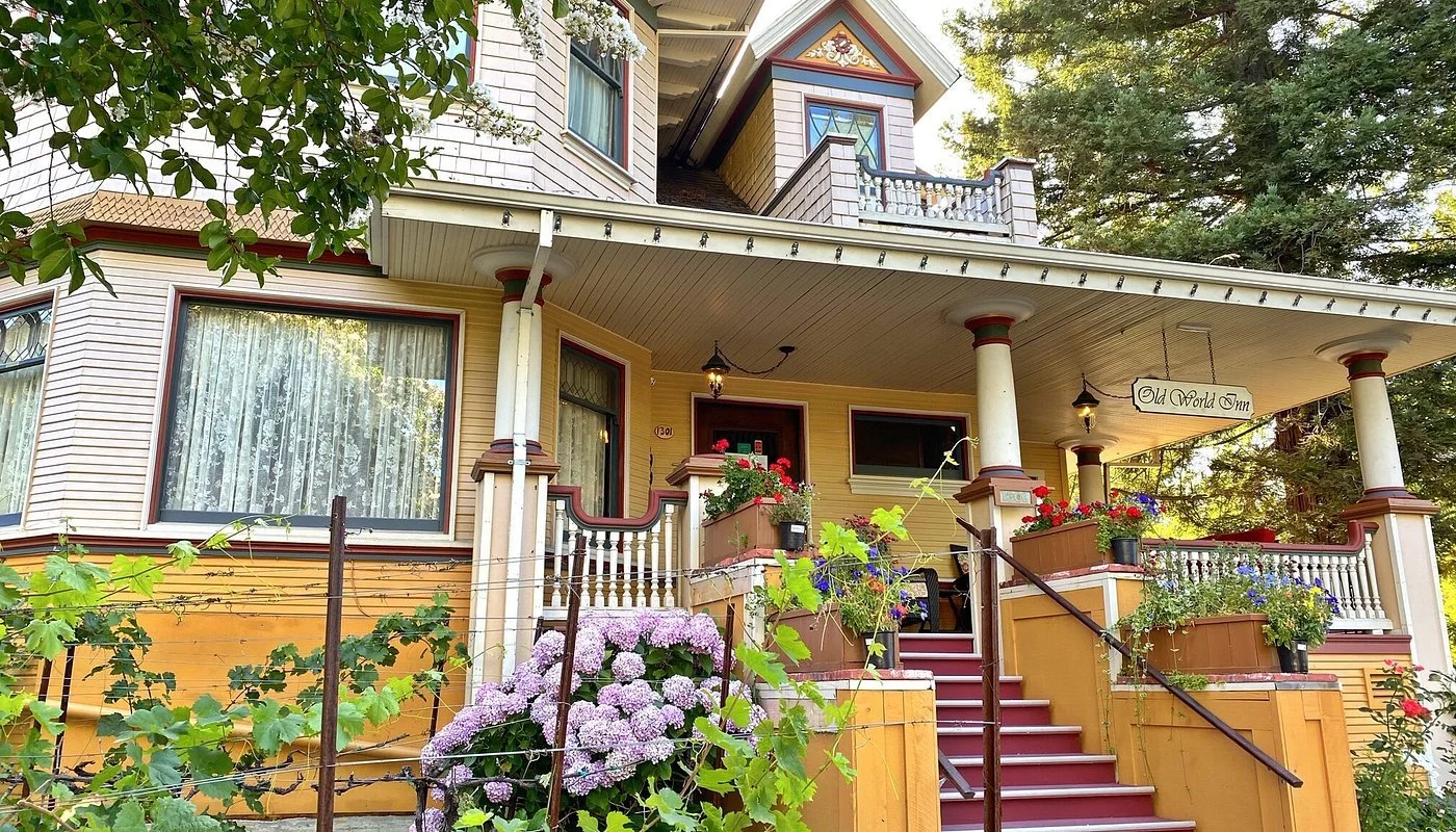 A yellow Victorian-style house with a porch decorated with flowers and plants, white columns, and a sign that reads 'Old World Inn.'