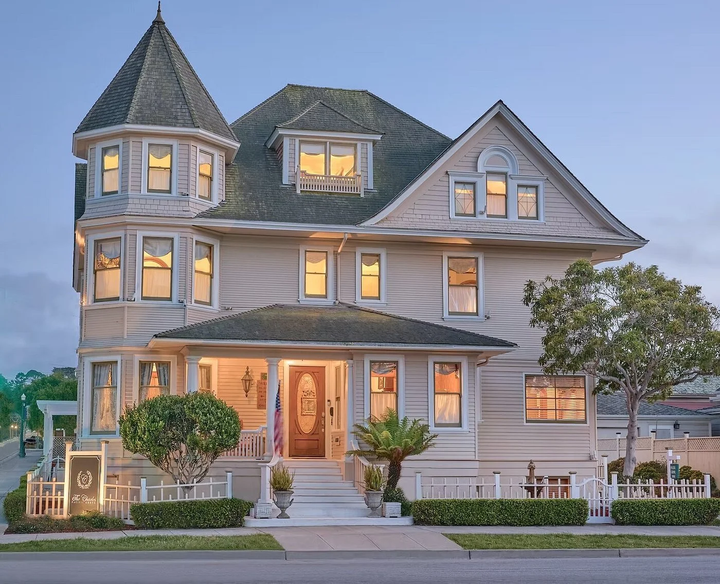 A large Victorian-style house with multiple stories, a turret, and peaked roof, lit from within, with white siding, front porch, stairs, and well-maintained garden, in the evening.
