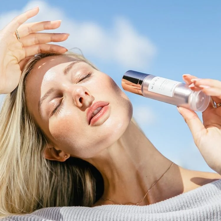 A woman with blonde hair outdoors on a sunny day, holding a bottle of skincare mist near her face with her eyes closed.
