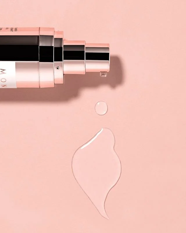 A pink and black bottle of skincare serum or moisturizer with a dropper applicator, lying on a pink surface with two drops of the product next to it.