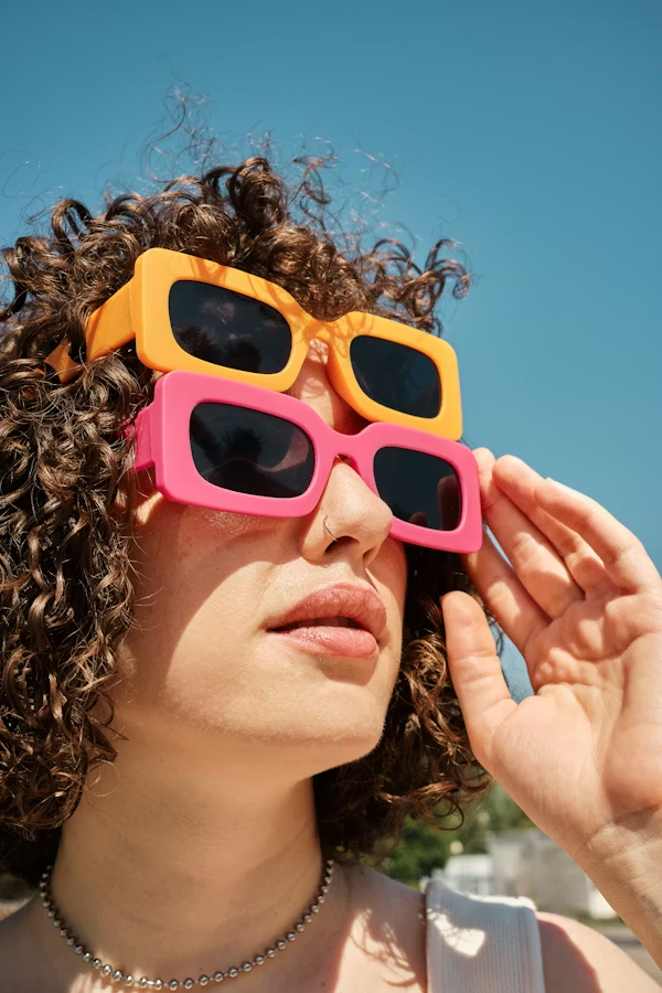 A woman with curly hair wearing oversized, layered sunglasses with yellow and pink frames against a clear blue sky.