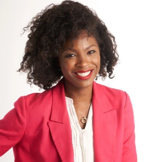 A smiling African American woman with curly hair wearing a pink blazer and white blouse, standing against a white background.