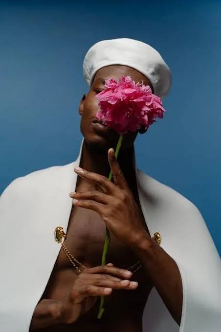 Person with dark skin wearing a white hat and white jacket, holding a pink flower in front of one eye, against a blue background.