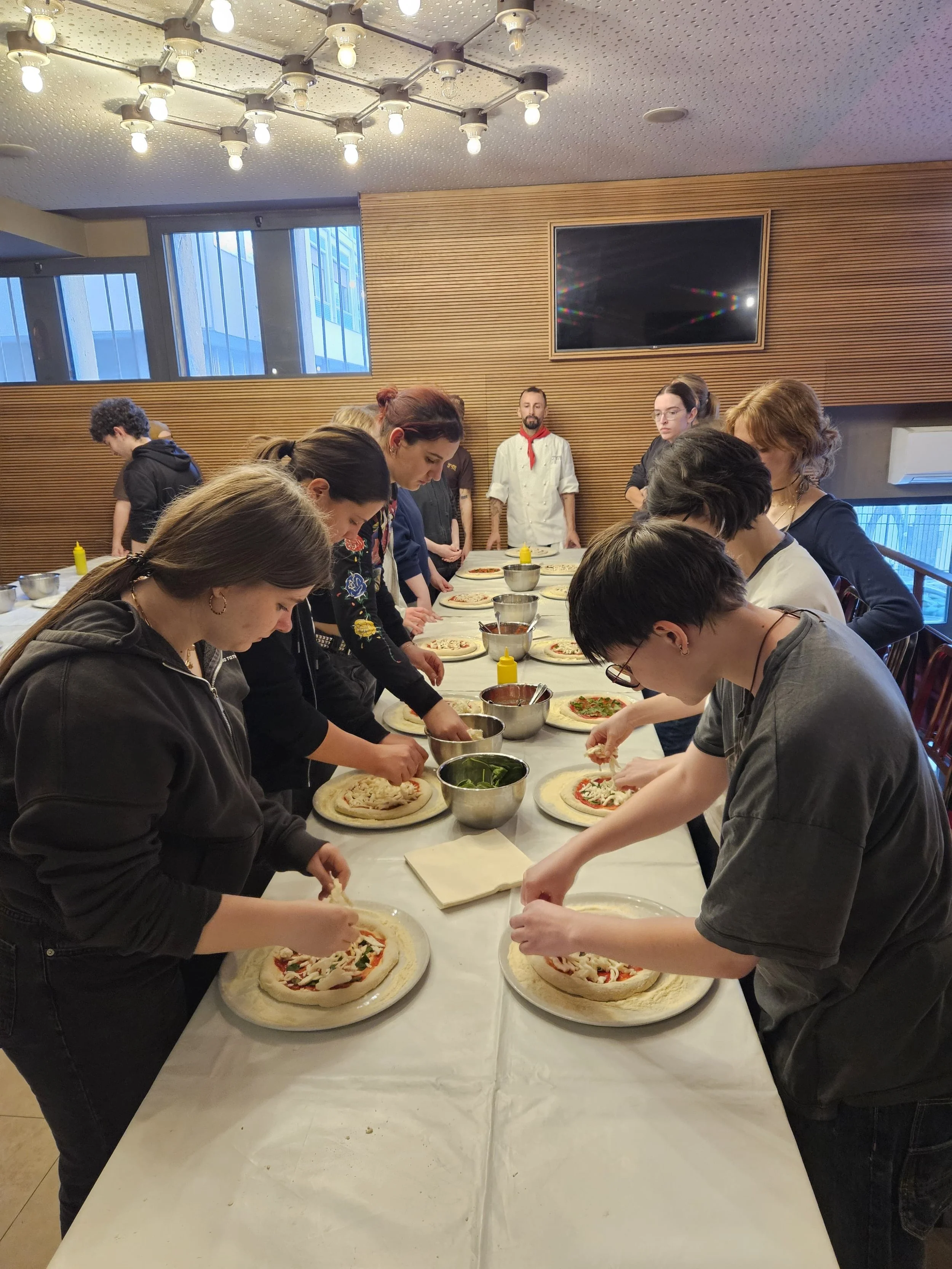 Menschen bereiten zusammen Pizzas auf einem langen Tisch in einer modernen, gut beleuchteten Küche zu.