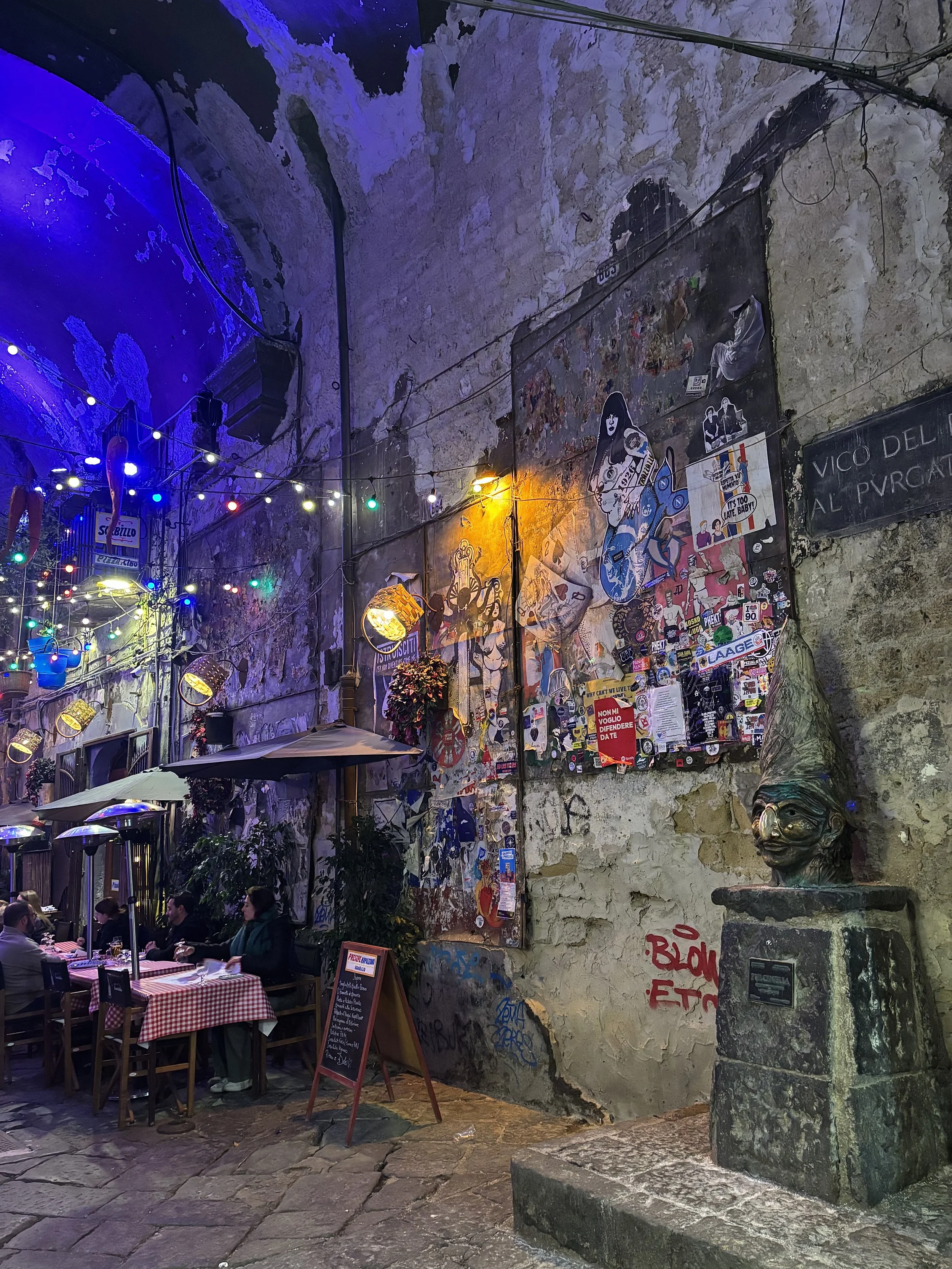 Eine Straßenszene vor einem italienischen Restaurant mit Tischen, die mit roten und weißen Tischdecken bedeckt sind, darüber hängen bunte Lichter. An der Wand sind Graffiti und Poster, und eine Skulptur eines Zwergkopfes auf einem Sockel steht rechts im Bild.