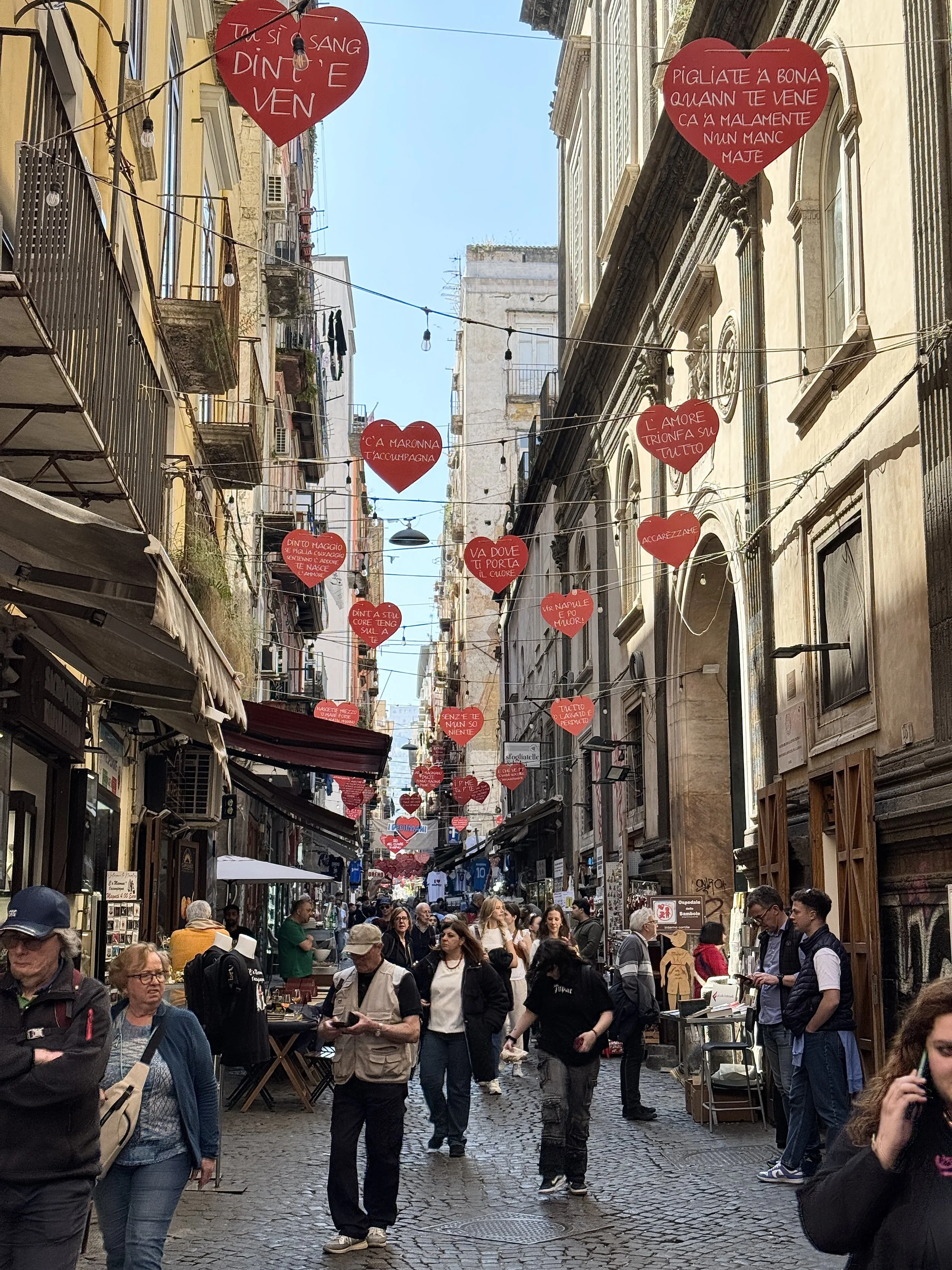 Straßenszene in einer belebten Einkaufsstraße mit roten Herzensdekorationen und Menschen beim Einkaufen und Flanieren.