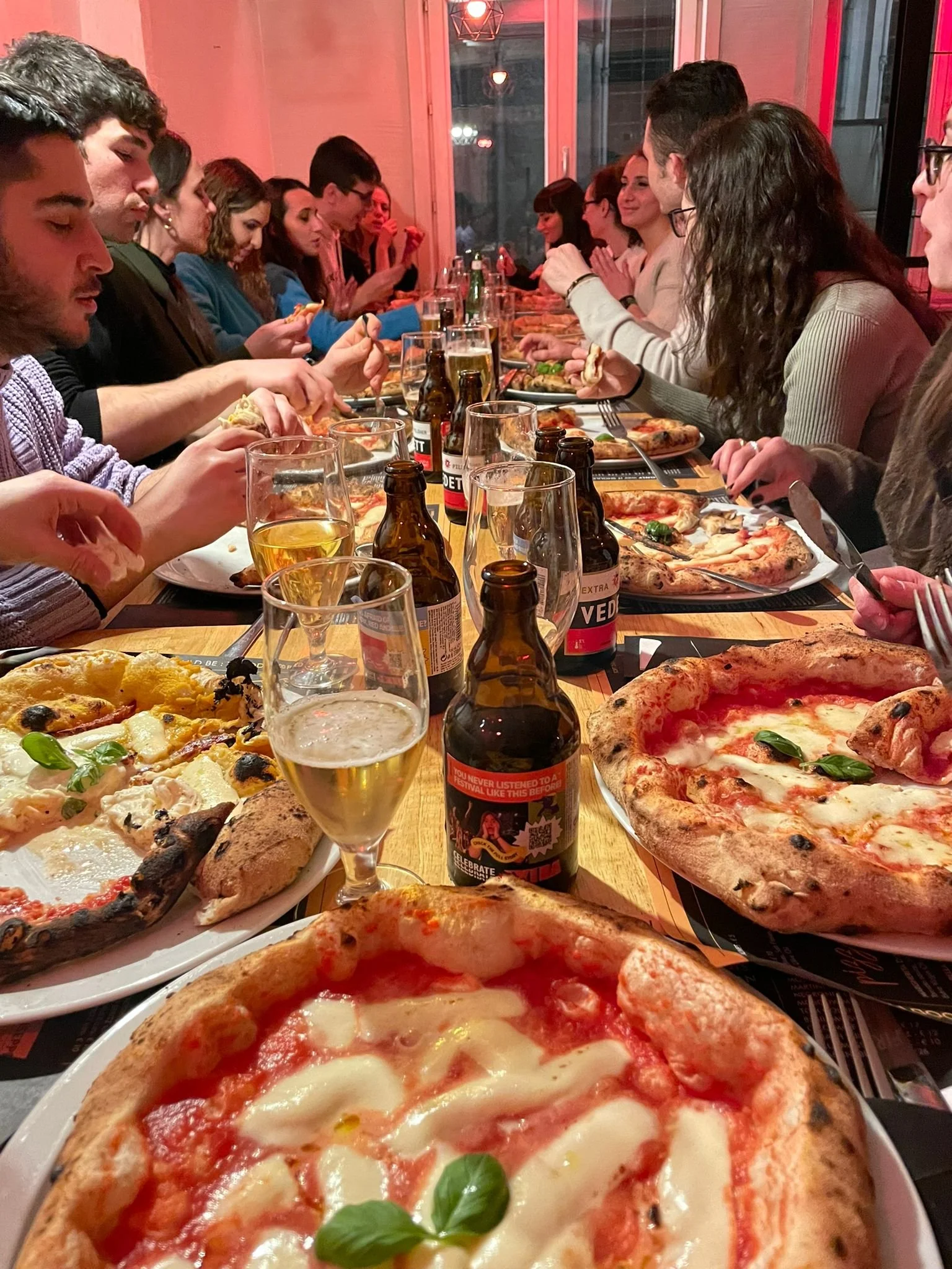Eine lange Tafel mit mehreren Personen, die Pizza essen und trinken, darunter Bier und Wein in einem Restaurant mit rotem Licht.