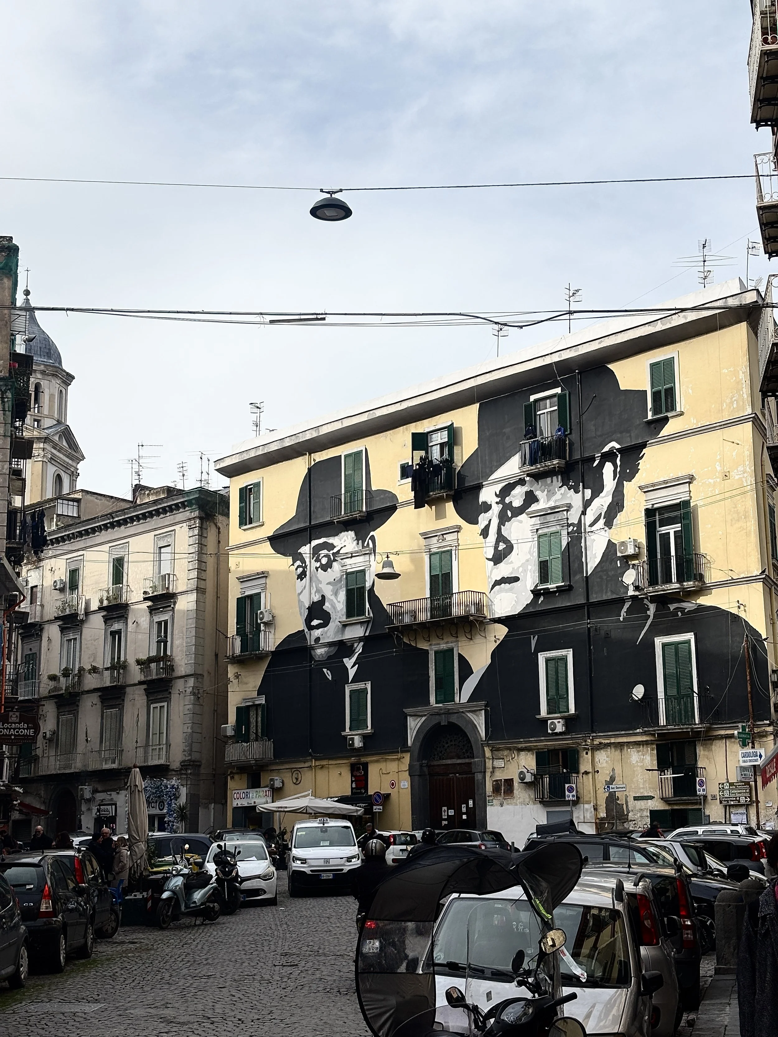 Straßenszene in Italien mit einem großen Wandgemälde von zwei Männern an einem Gebäude, umgeben von parkenden Autos und Motorrollern.