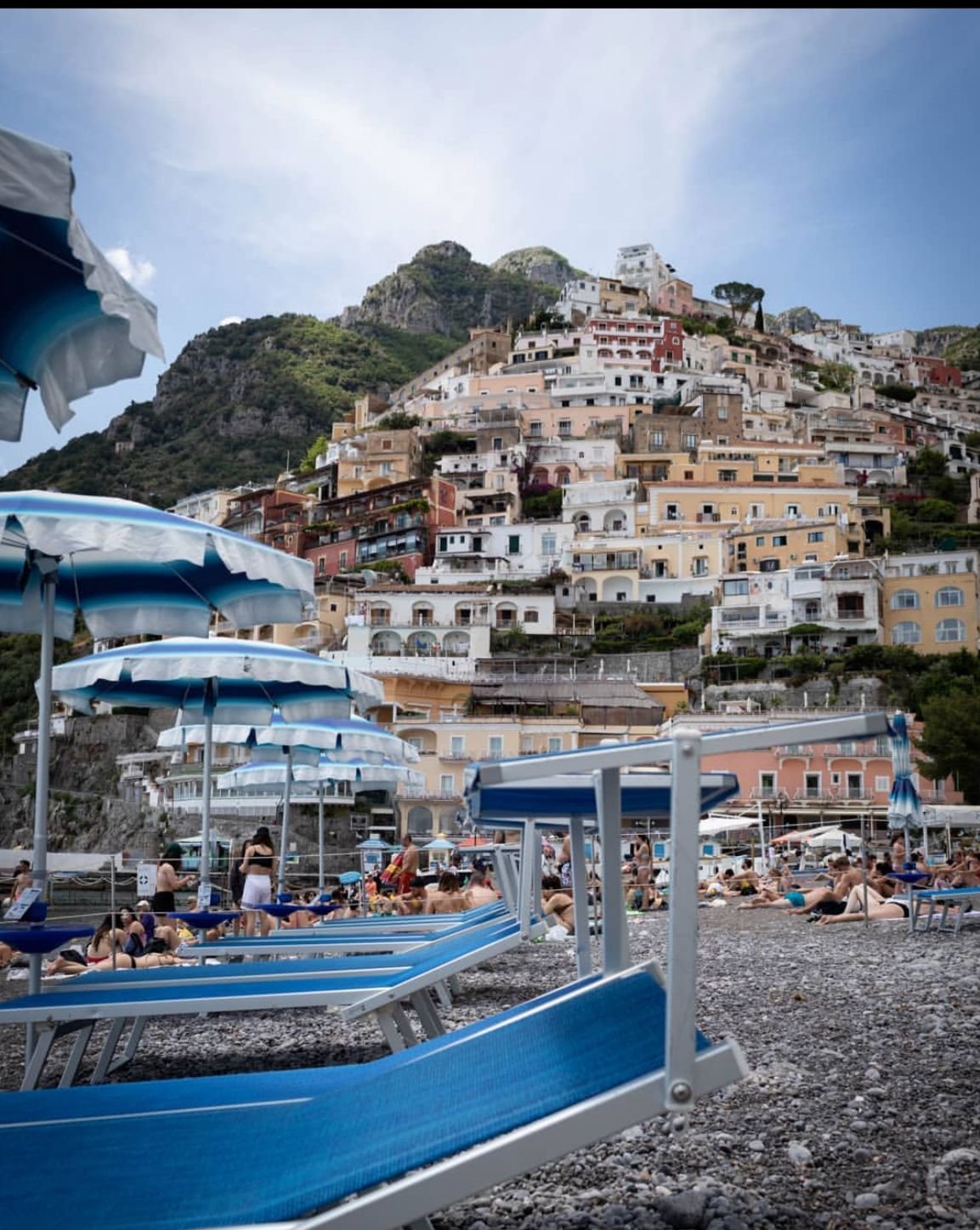 Strand mit Sonnenschirmen und Sonnenliegen, im Hintergrund bunte Häuser auf einem Hügel an der Küste.