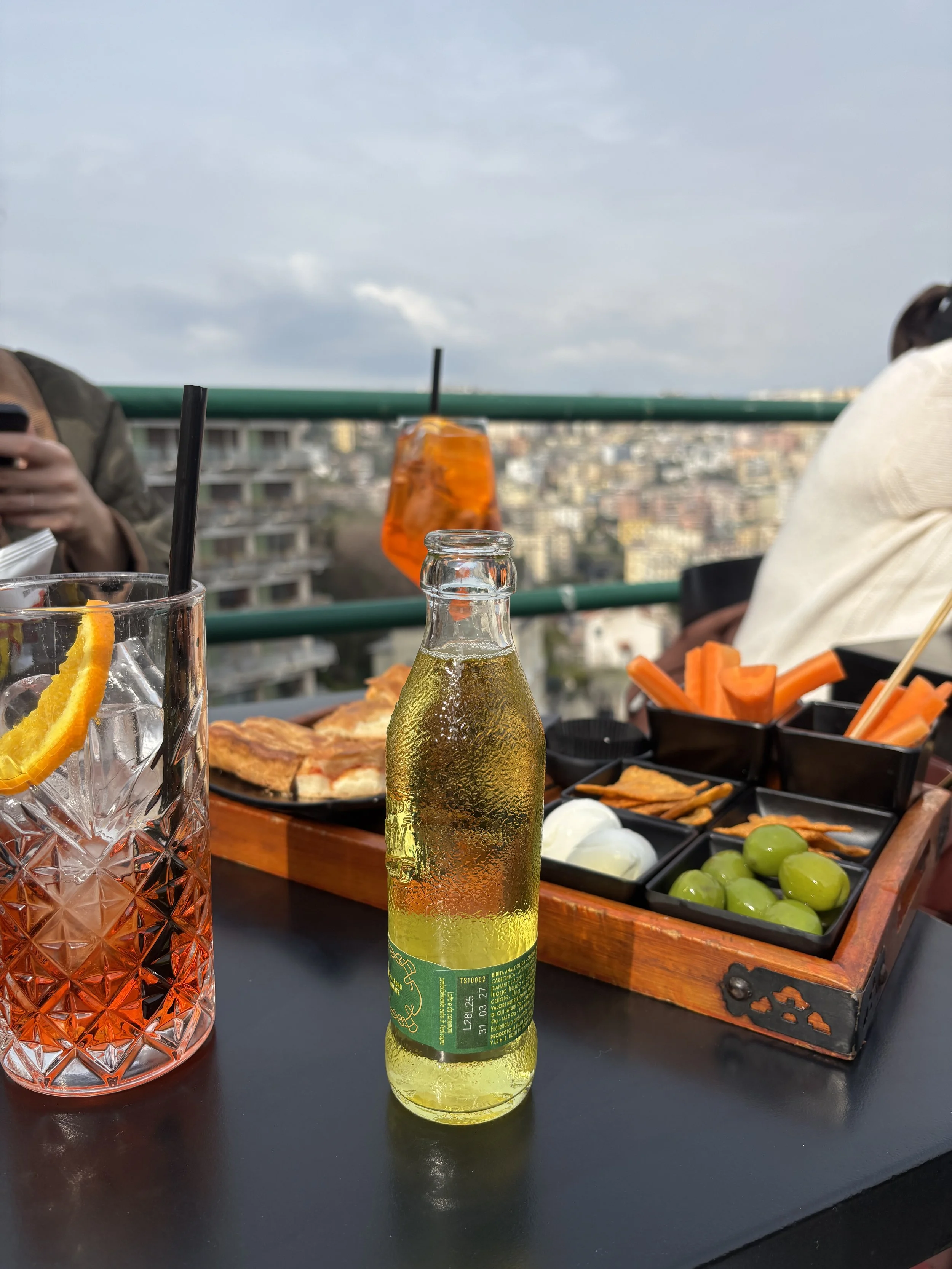Getränk mit Eiswürfeln und Zitronenscheibe, klassische Cocktailglas, Flasche mit Glasur, Snackplatte mit grünem und gelbem Gemüse, am Tisch auf einer Terrasse bei wolkigem Himmel in einer Stadt.