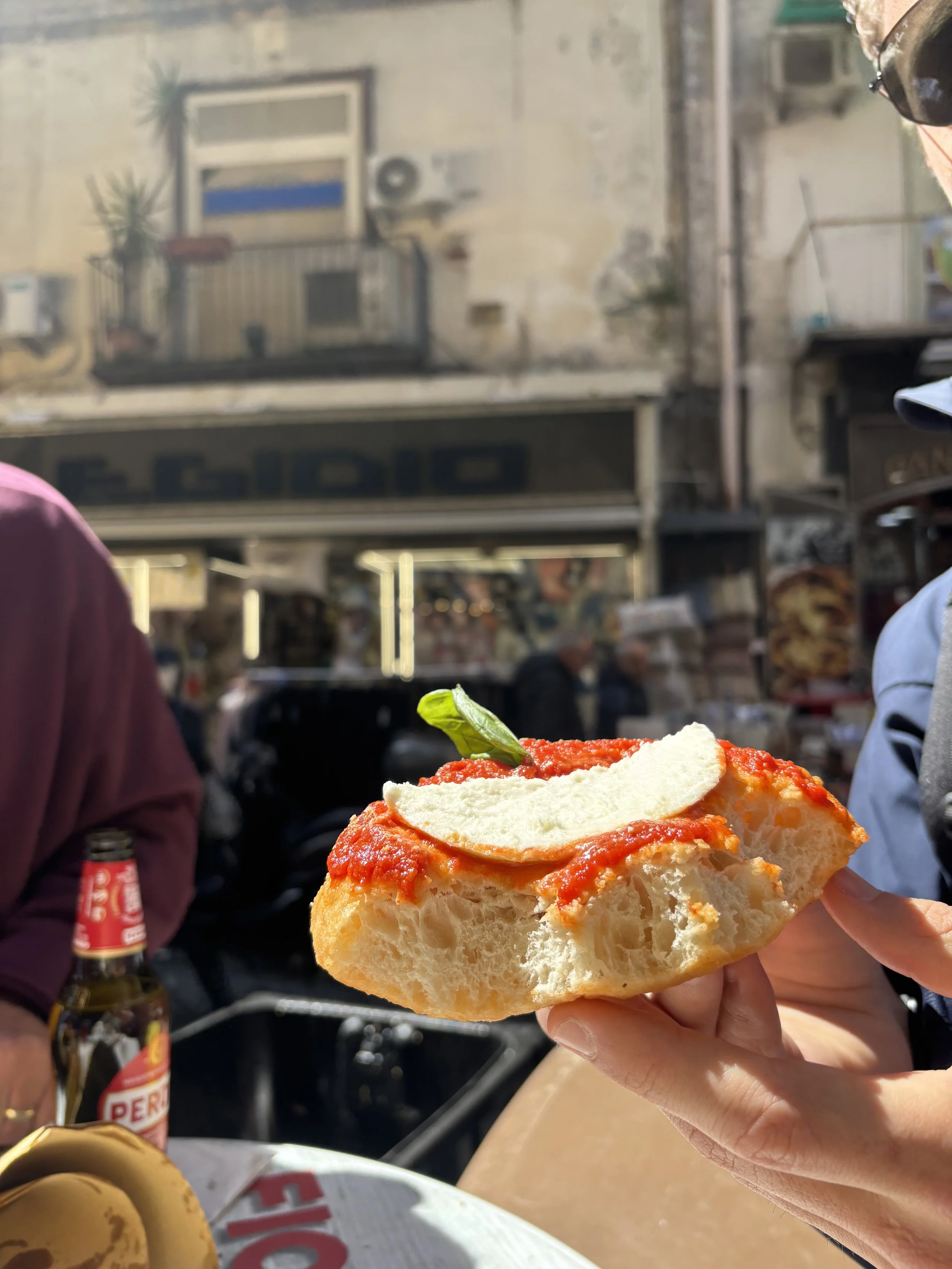 Nahaufnahme einer Person, die ein Stück Pizza mit Tomatensauce, Mozzarella und Basilikum hält, im Freien auf einer Straße in Italien.