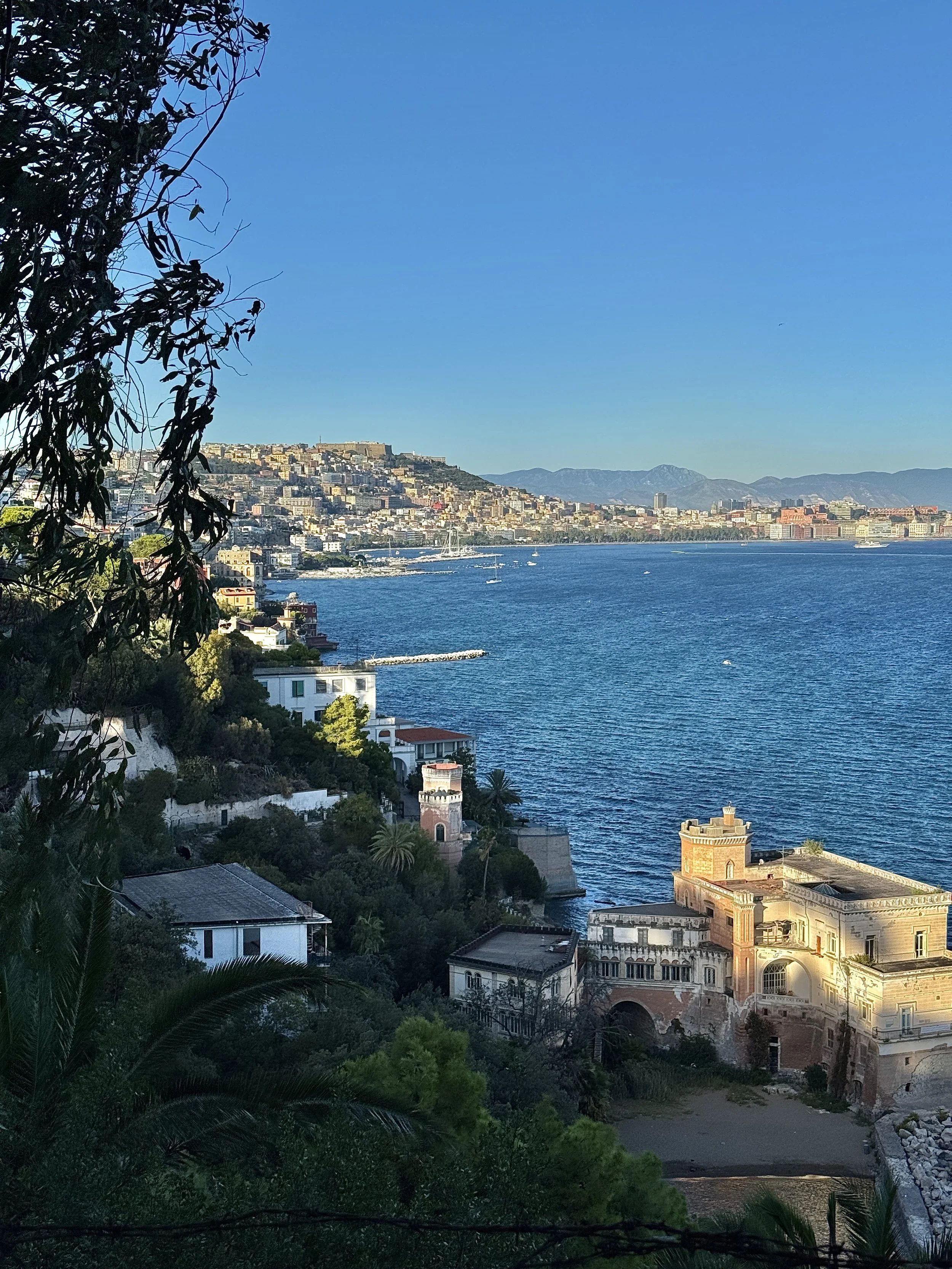 Blick auf eine Küstenstadt mit alten Gebäuden, Wasser und Bergen im Hintergrund, unter einem blauen Himmel.
